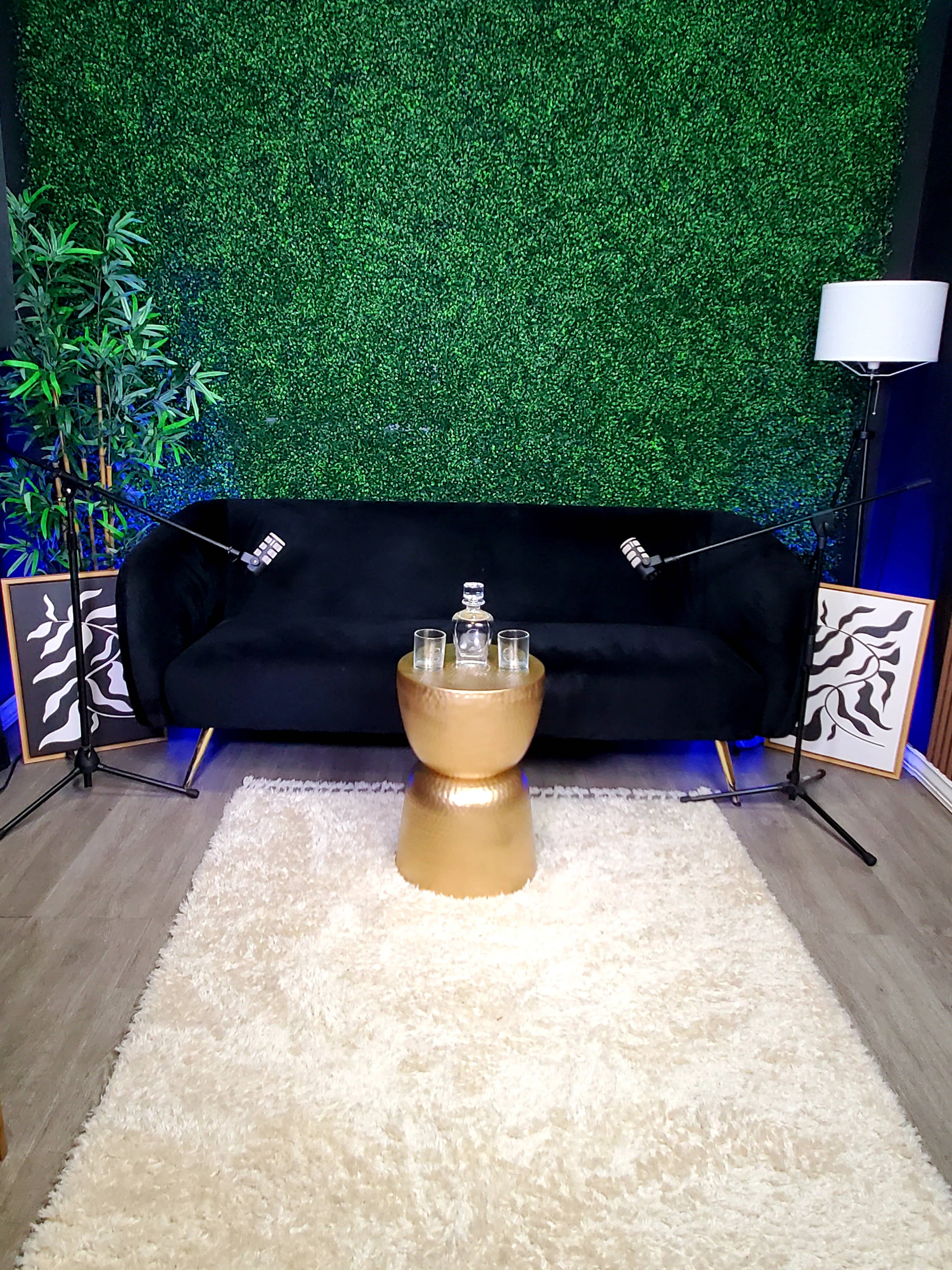 A black sofa sits against a green foliage backdrop, with a gold table holding glasses at its center, flanked by lamps and framed art on the floor.