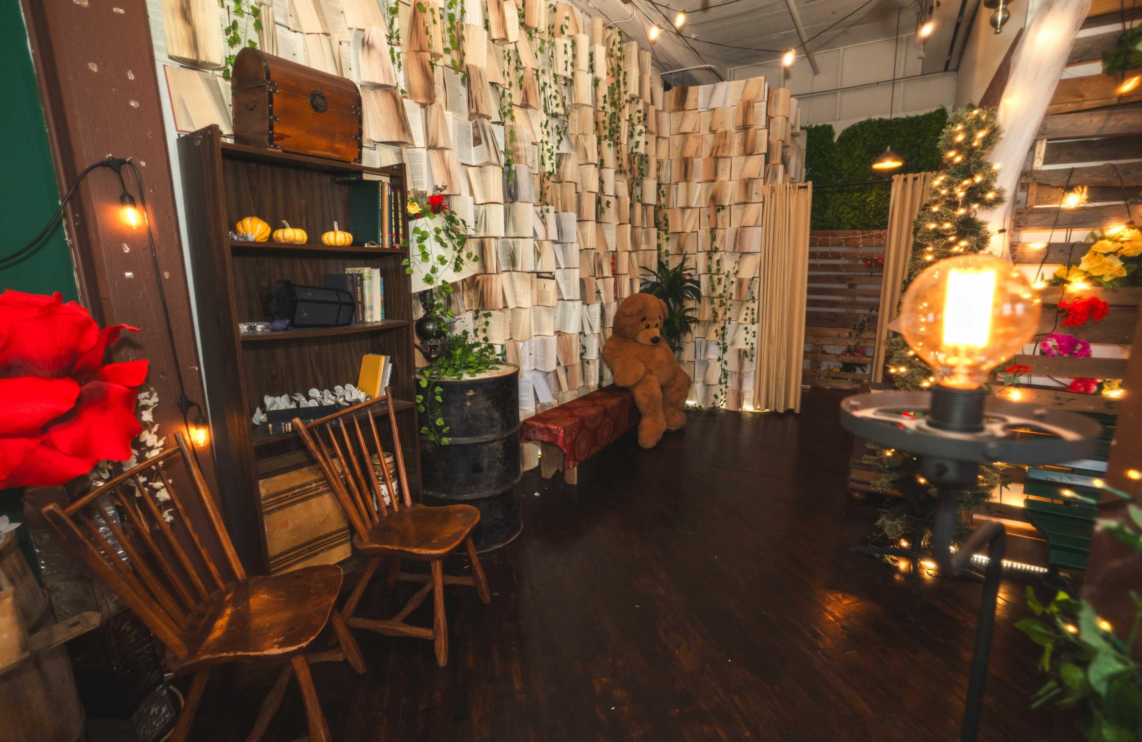 The scene features a cozy room with a book-themed wall made of stacked pages, a large teddy bear sitting on a bench, two wooden chairs, and warm string lights illuminating the space.