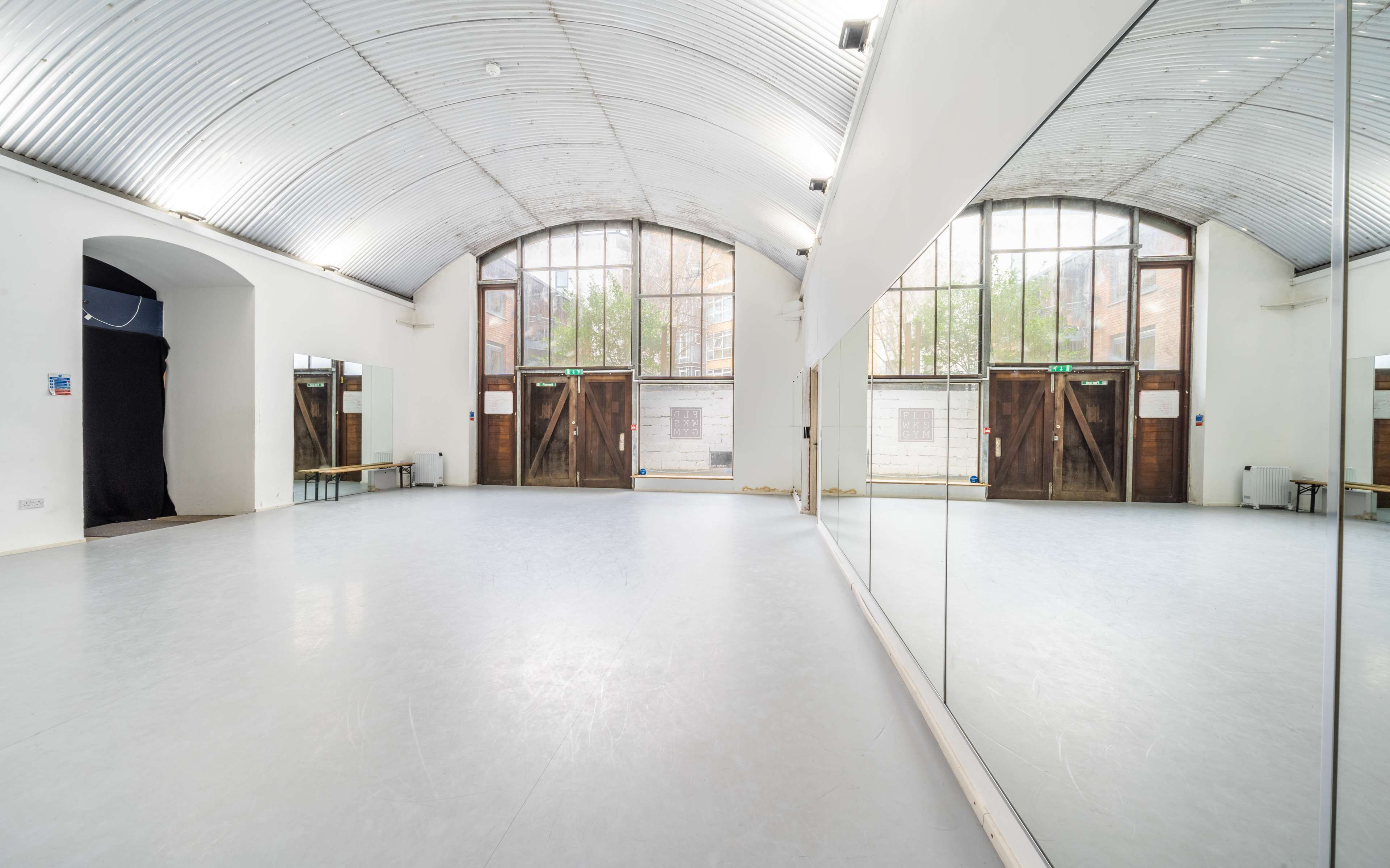 A spacious, well-lit dance studio featuring large mirrors, a high arched ceiling, and wooden double doors with glass panels.