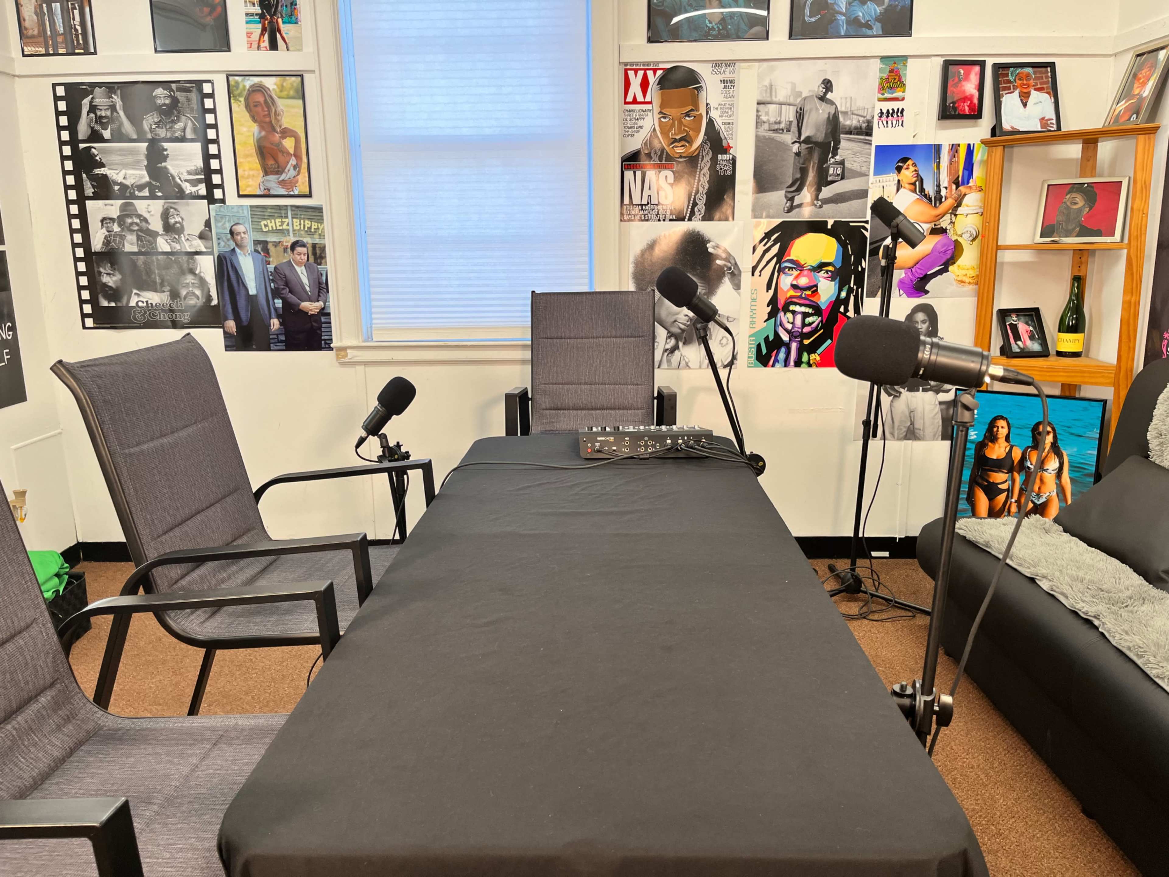 A podcast recording studio with a table, three chairs, and microphones setup against a wall adorned with various music posters.