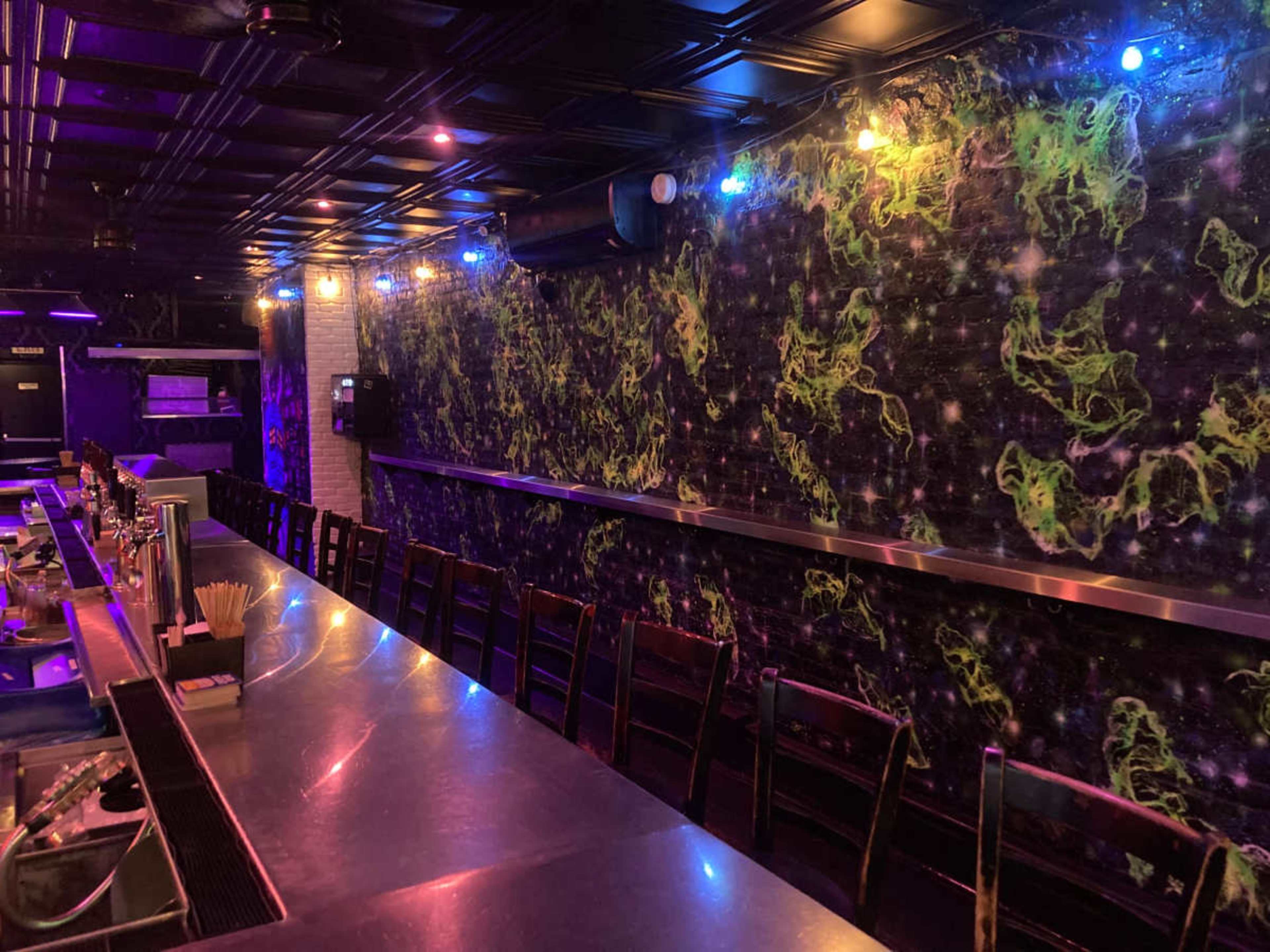A dimly lit bar with a metal countertop, wooden chairs, and a wall featuring colorful, abstract designs illuminated by vibrant lights.