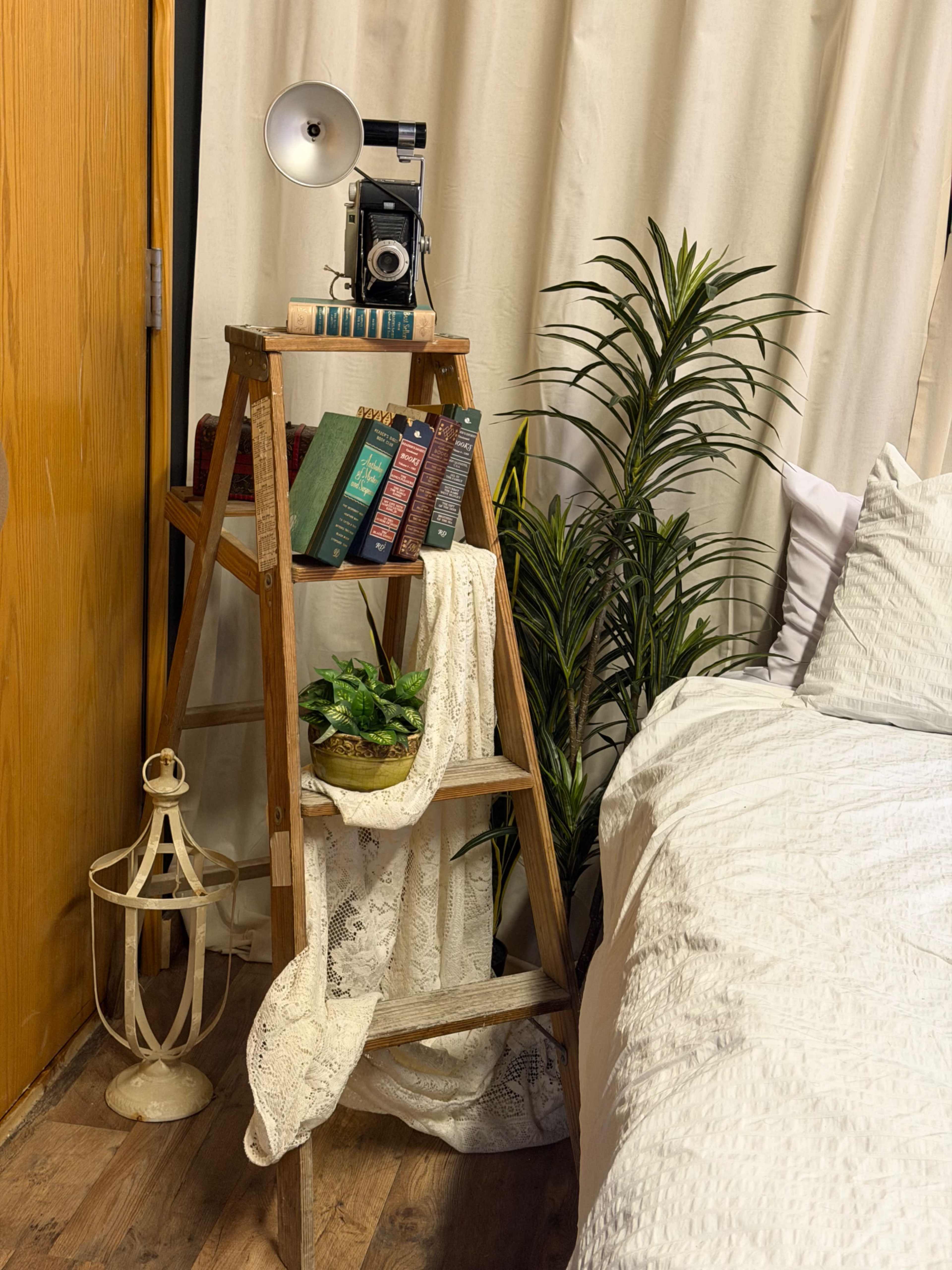 A wooden ladder shelf holds books and a vintage camera, while a potted plant and decorative items are placed beside a bed with a white quilt.