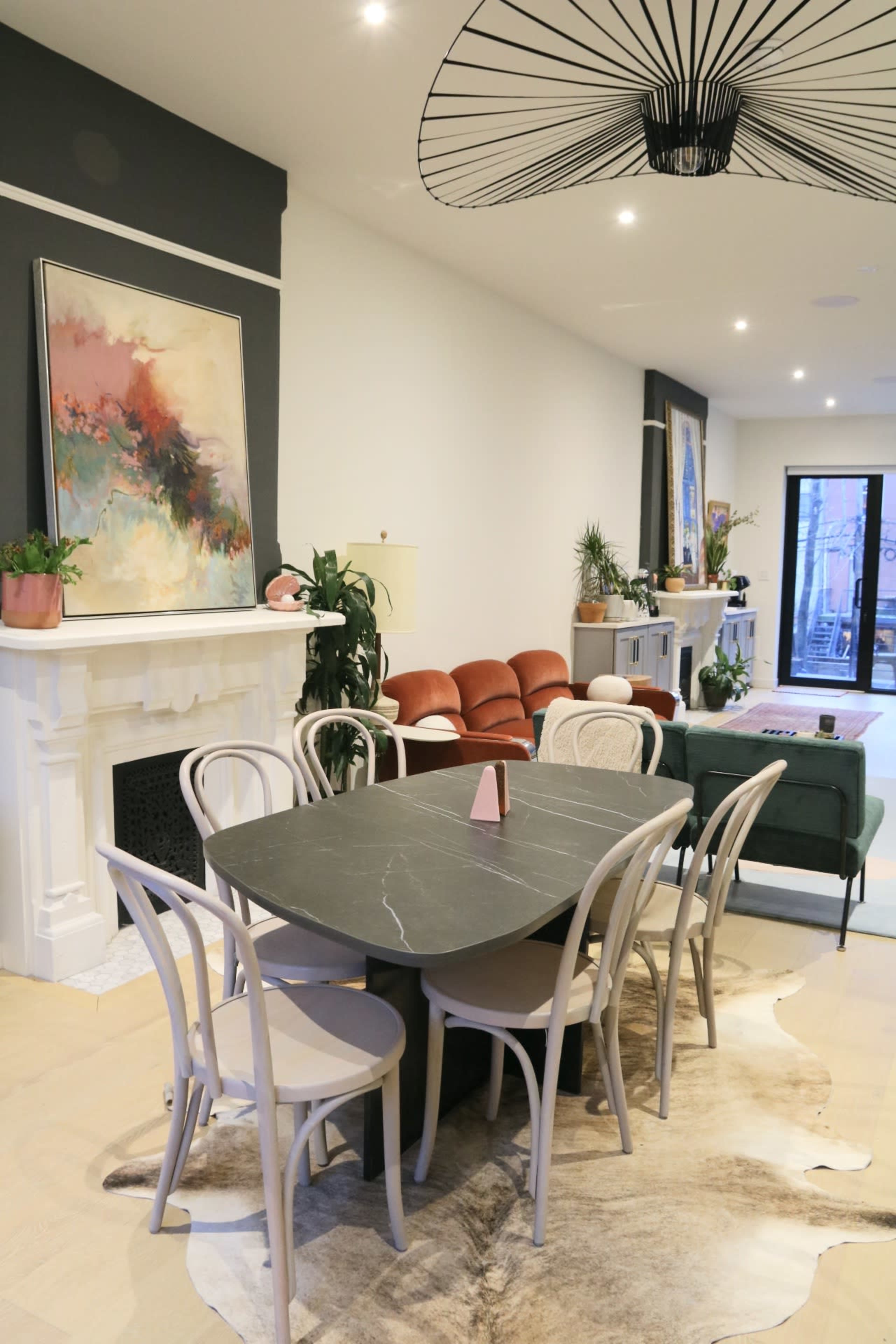 The image shows a modern dining area with a black marble table surrounded by white chairs, a decorative area rug, and a large artwork on the wall, adjacent to a living space with a green sofa and red chairs.