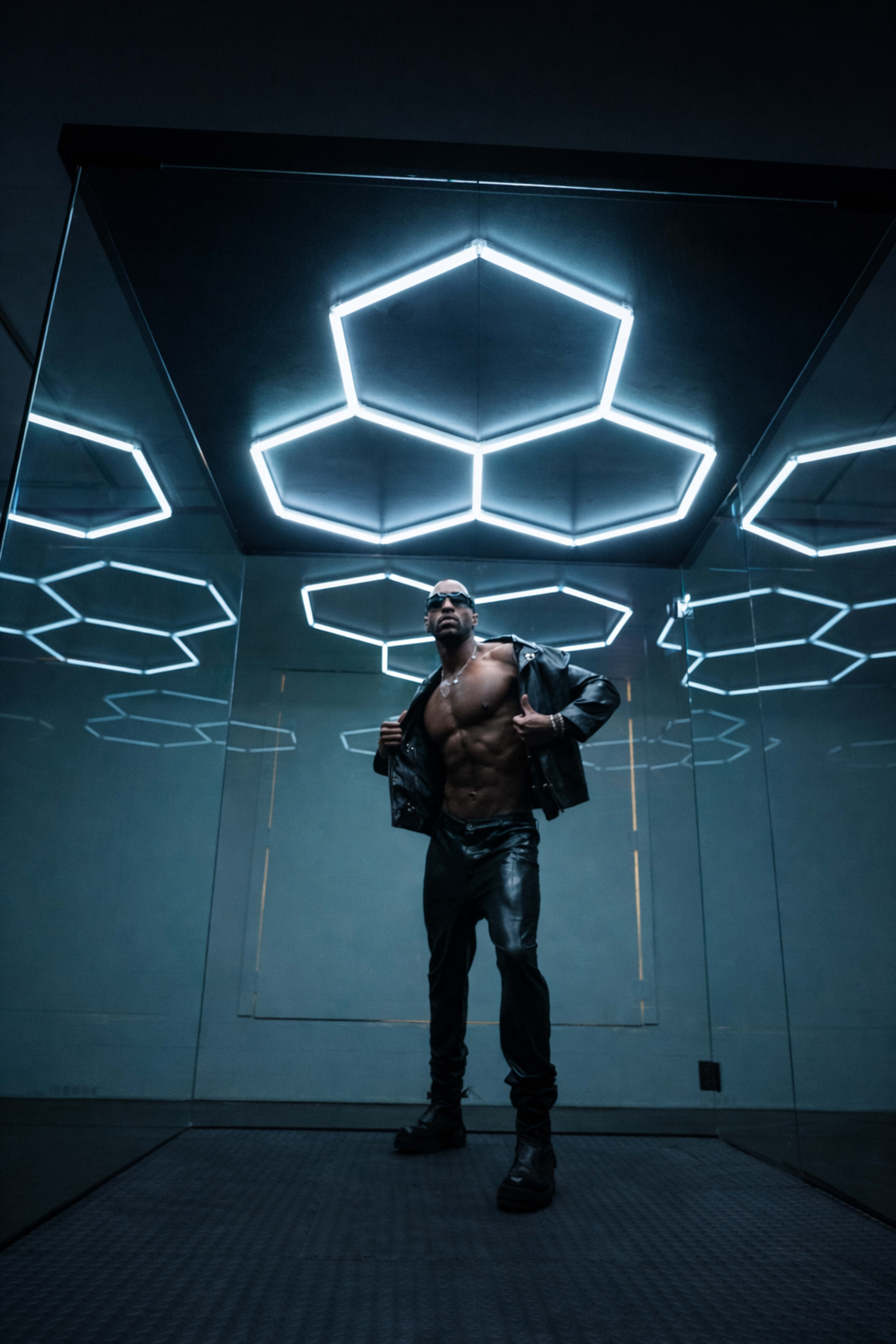A muscular person in a black leather jacket stands confidently under a geometric light fixture in a mirrored room.