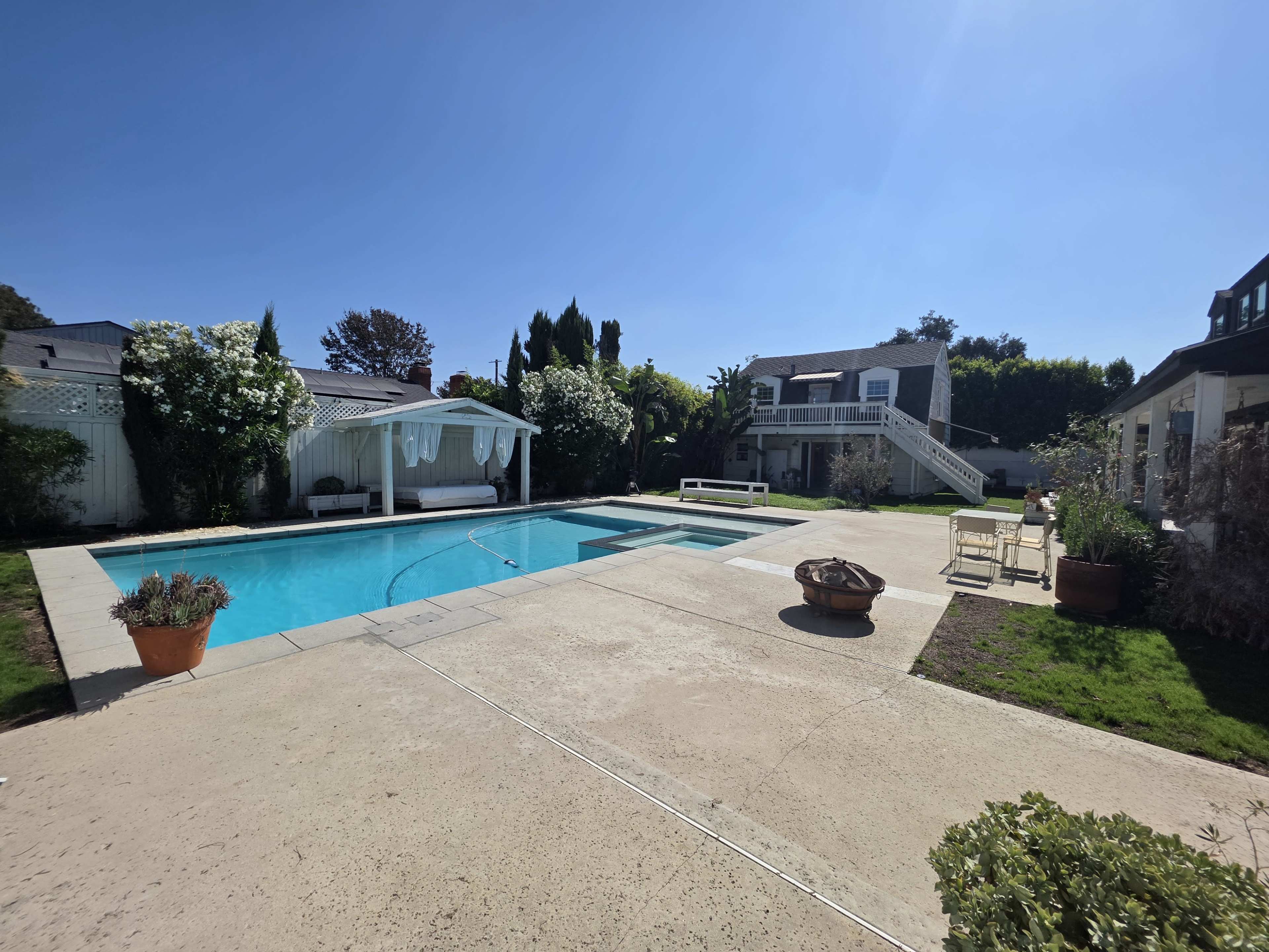 The image shows a sunny backyard with a swimming pool surrounded by a concrete patio, lounge chairs, potted plants, and a house with a second story in the background.