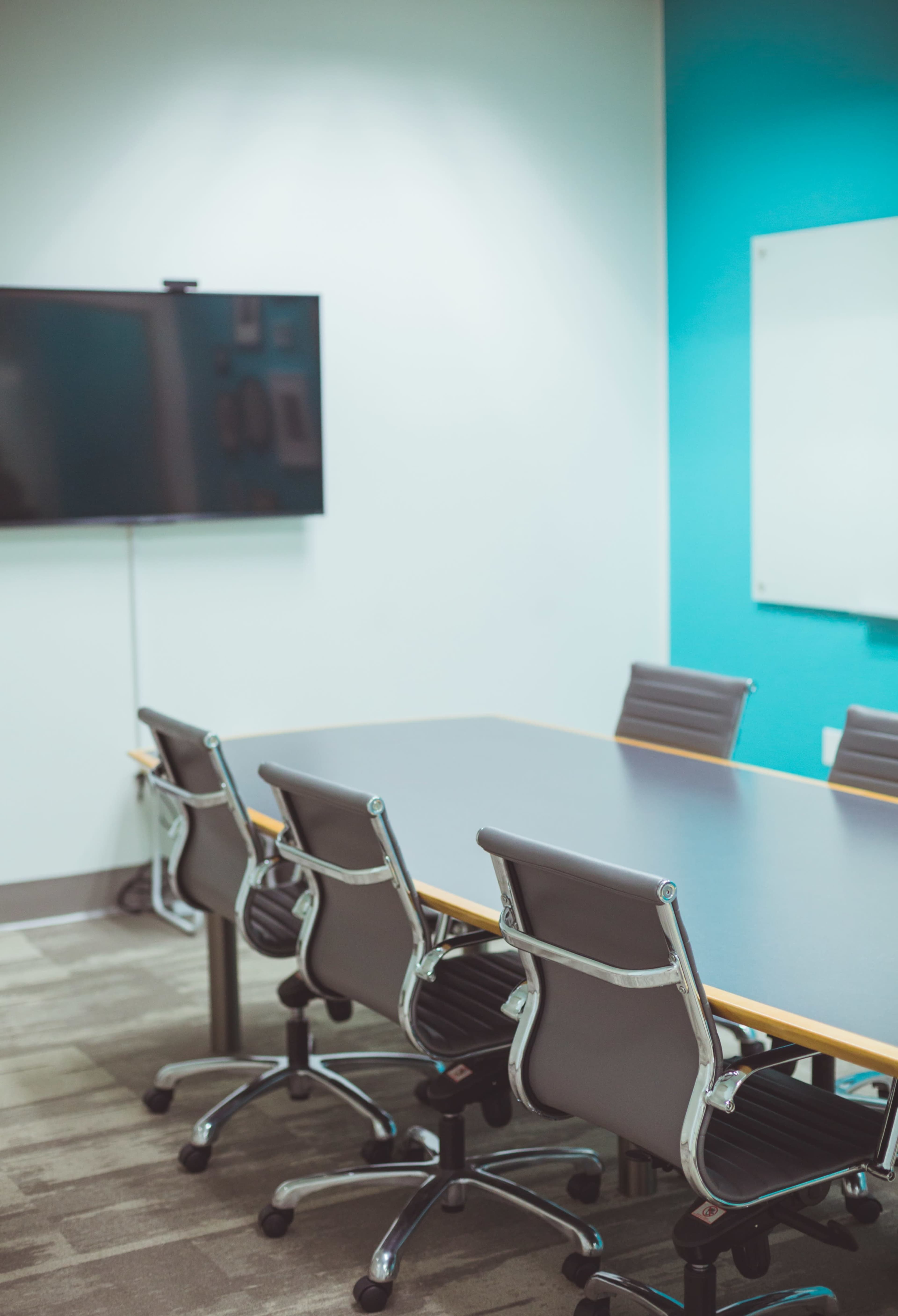A conference room features a large table surrounded by gray rolling chairs, with a television mounted on the wall and a whiteboard nearby.