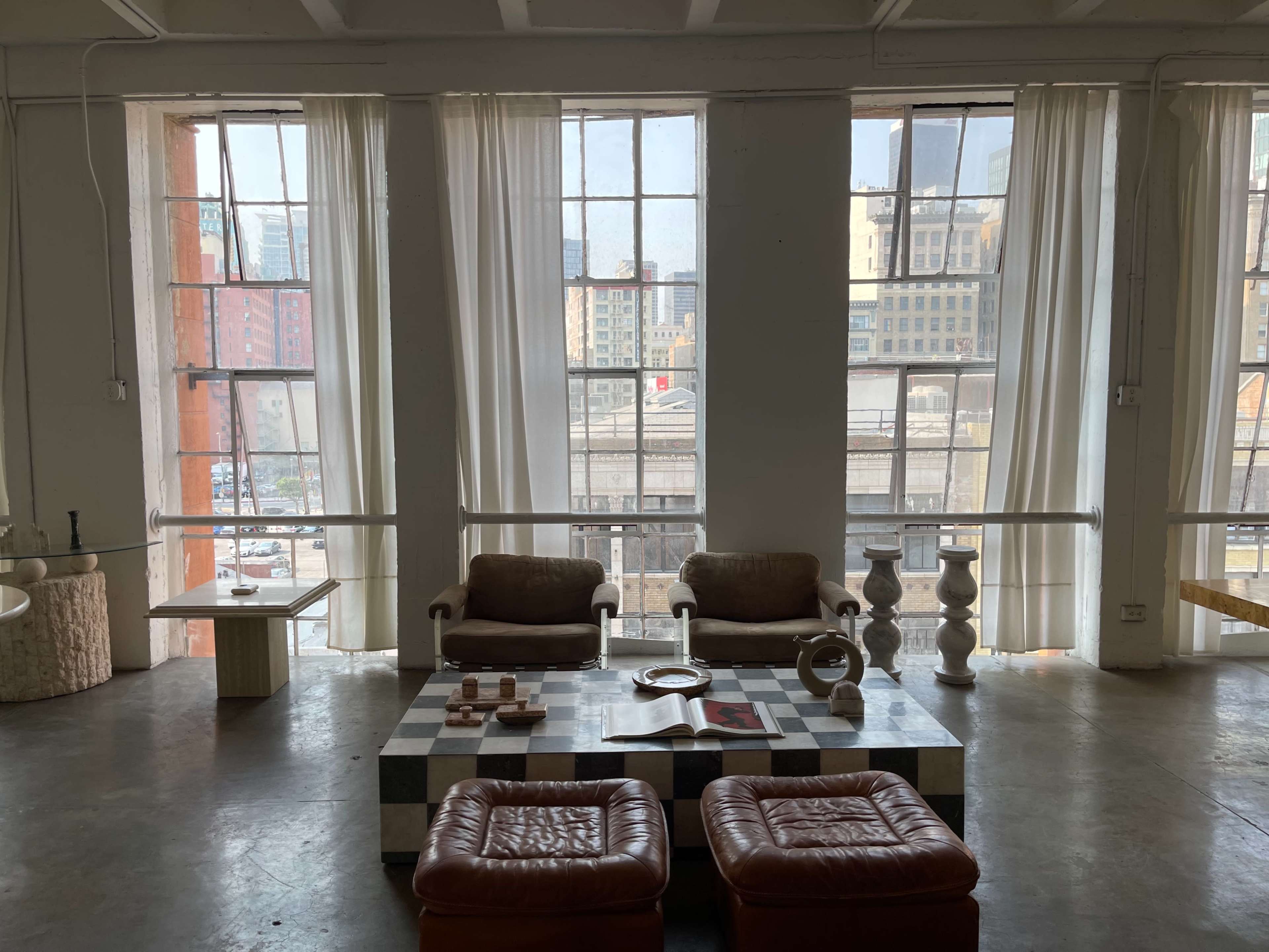 A minimalist living space with two brown chairs and a checkered coffee table, surrounded by large windows that let in natural light.