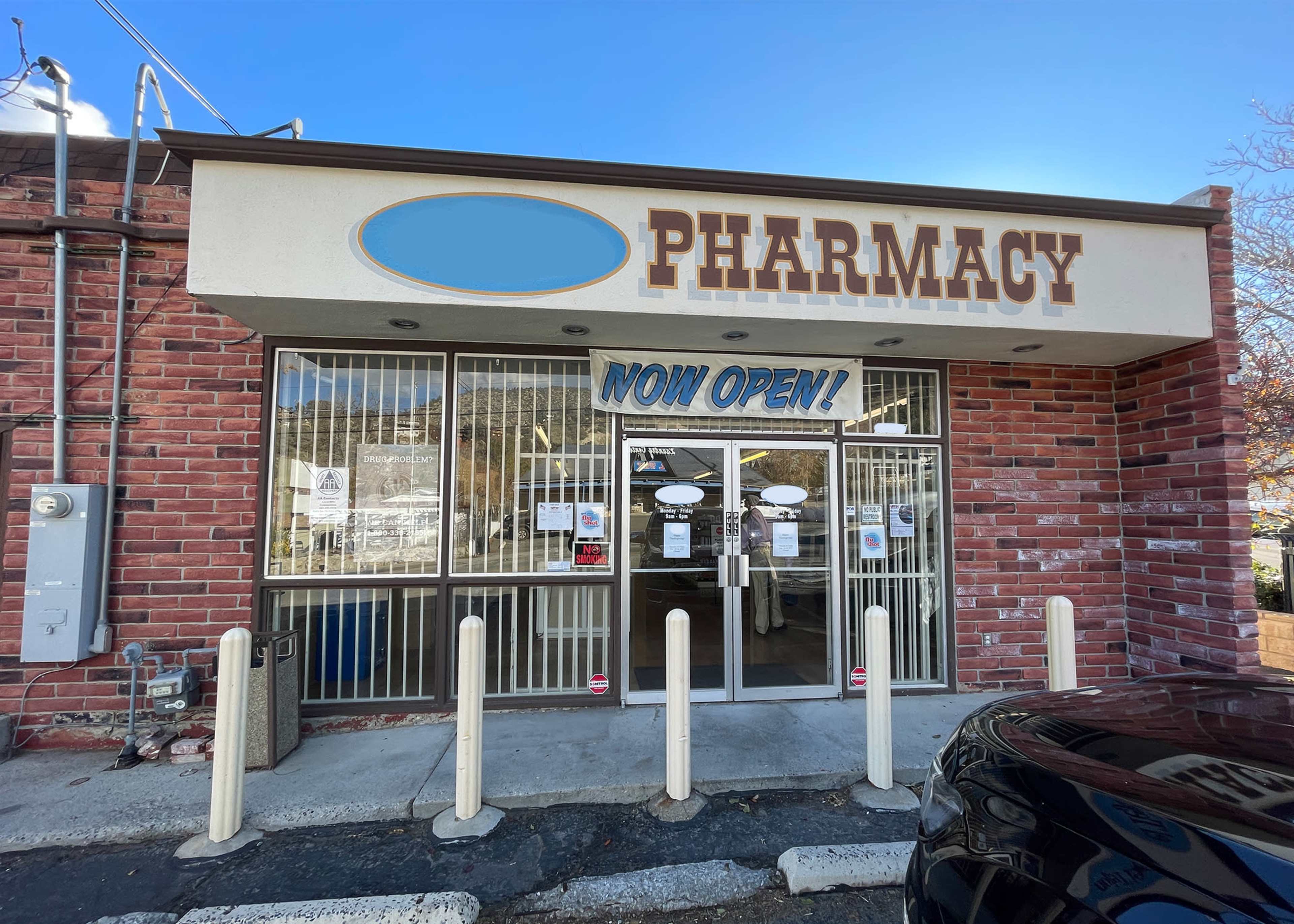 A pharmacy storefront with a large "NOW OPEN!" sign displayed in the front window.