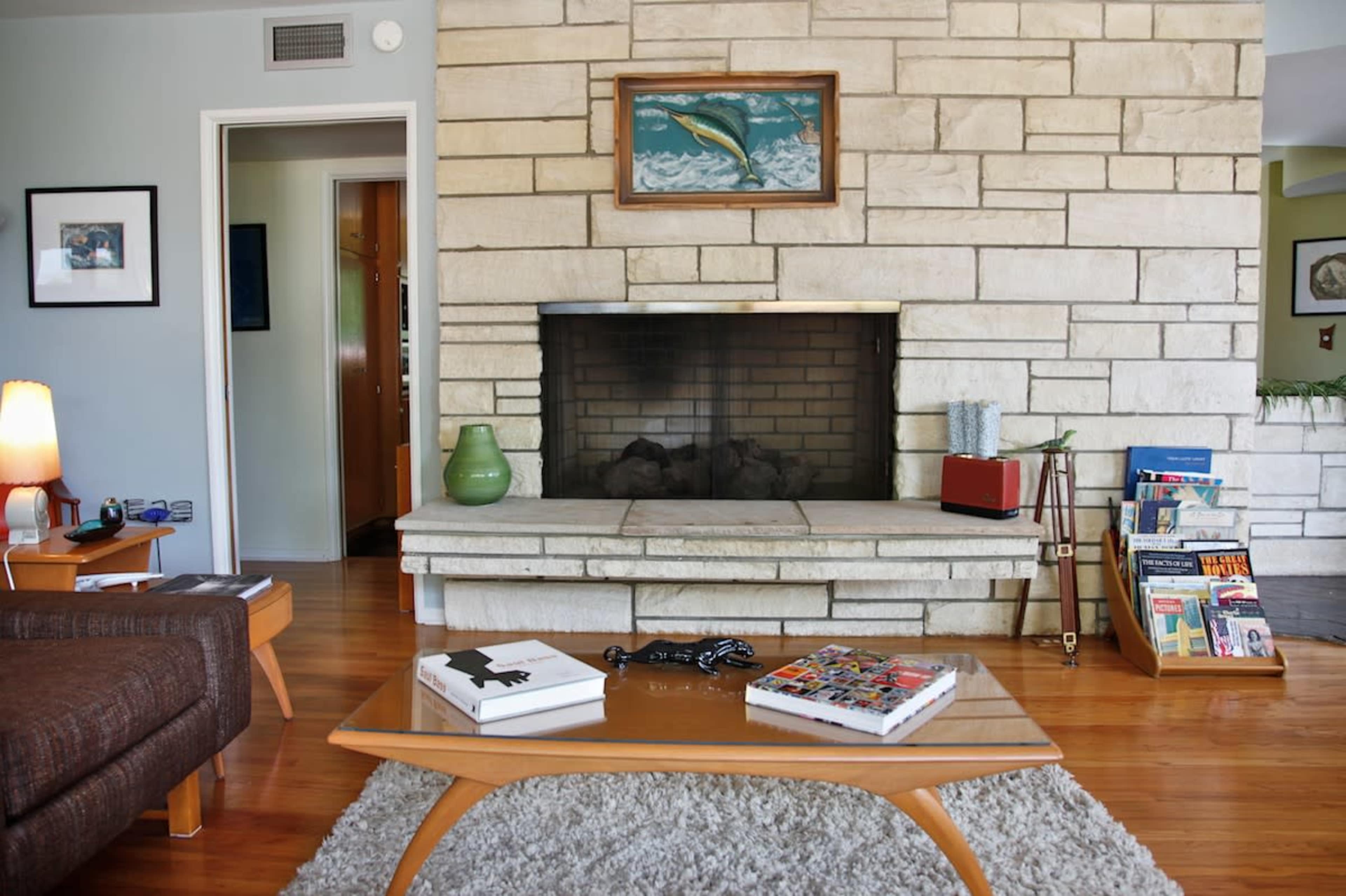 A cozy living room features a stone fireplace, a wooden coffee table with books on it, and a green vase on the hearth.