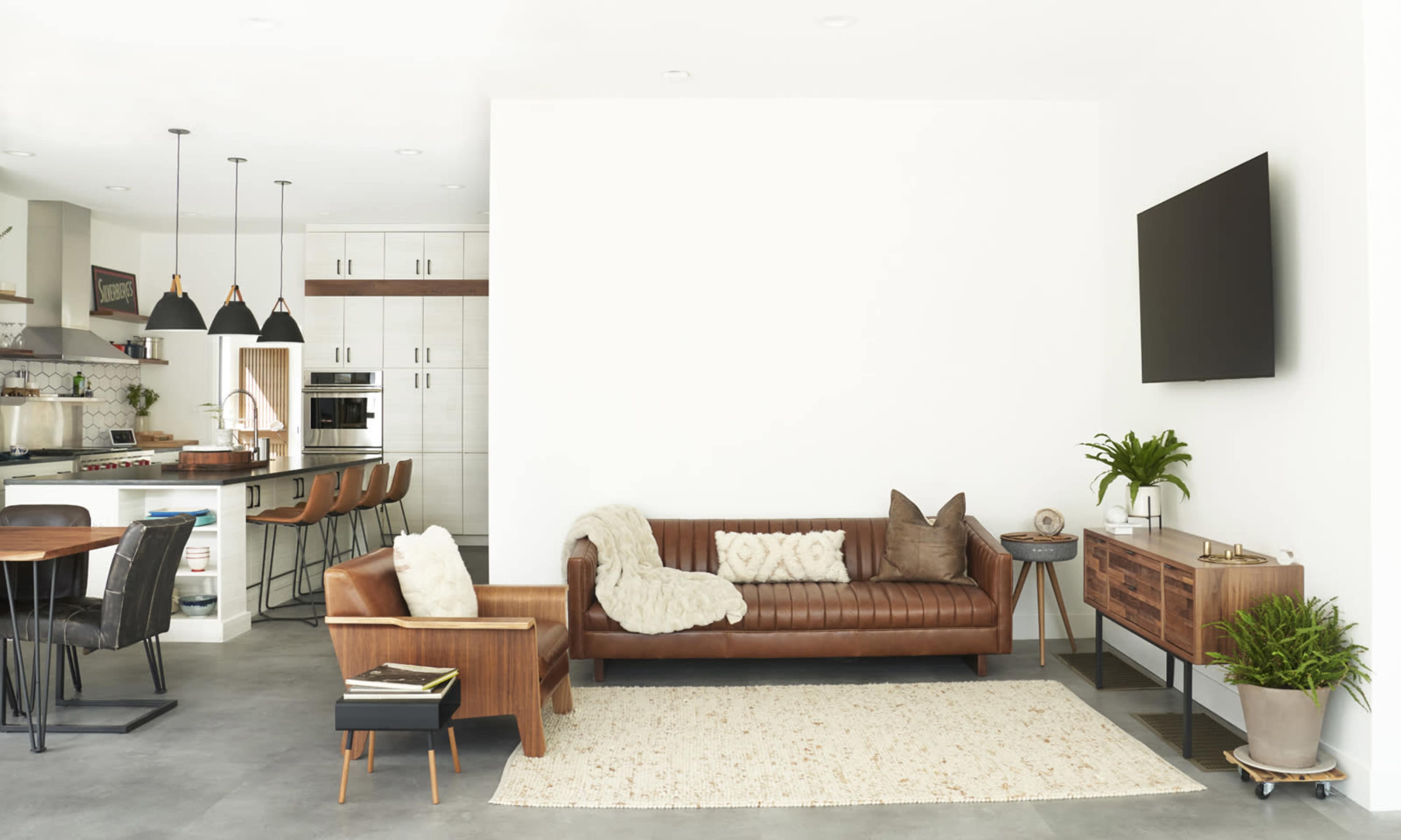 A modern living room features a brown leather sofa, a wooden armchair, a white wall, and a flat-screen TV, with an open kitchen and dining area in the background.