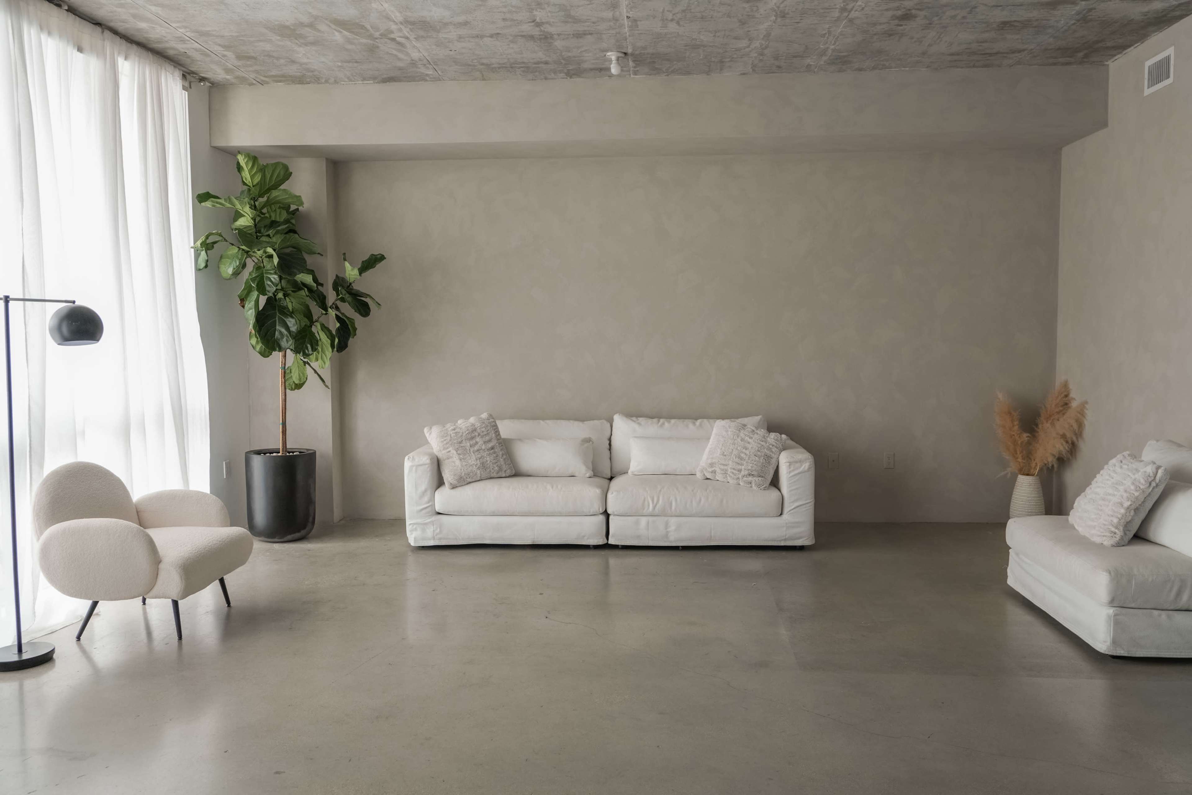 A minimalist living room features two white sofas, a floor lamp, and a large potted plant, with light-colored walls and a concrete floor.