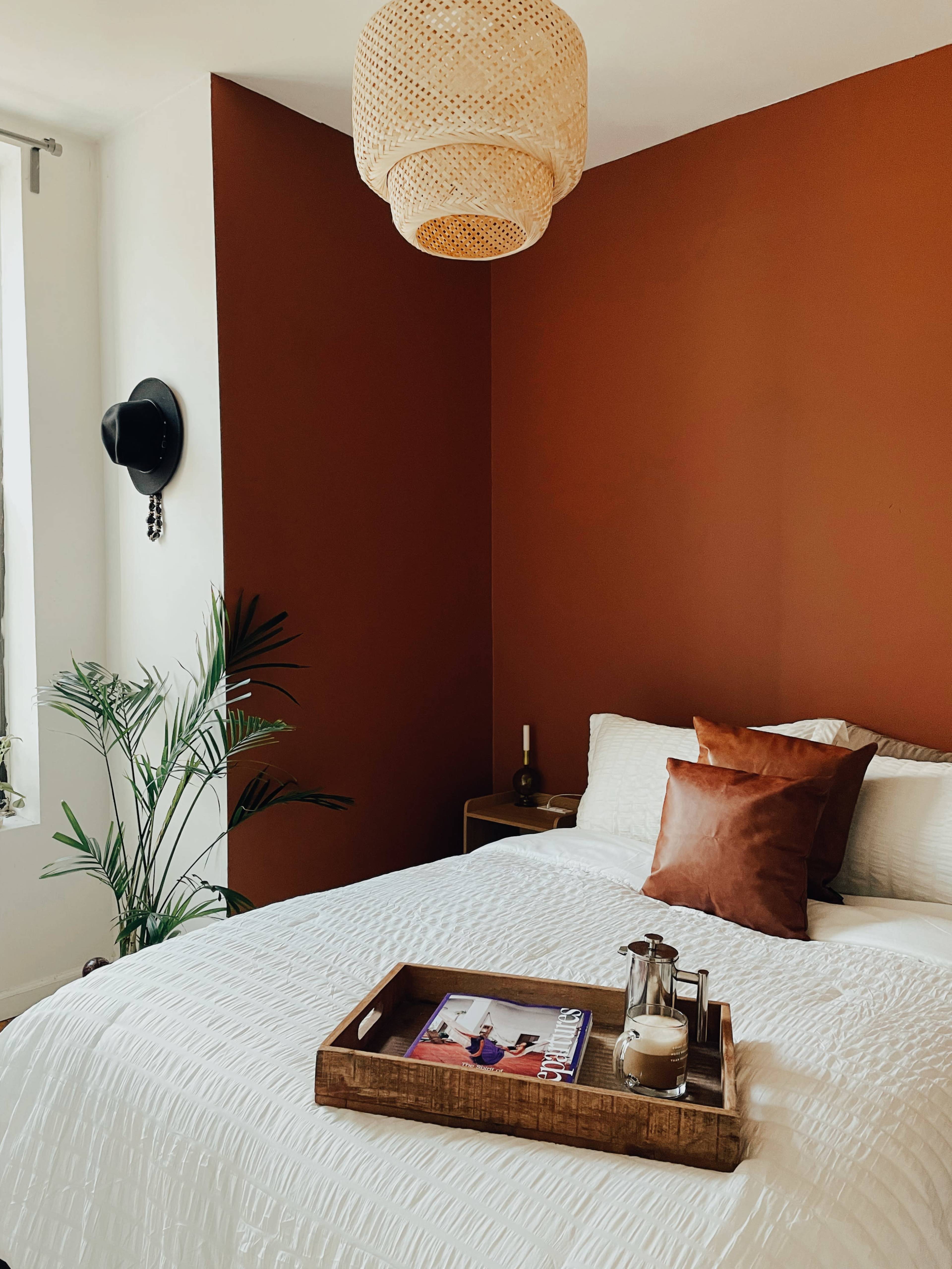 A bedroom features a white bed with a brown pillow and a wooden tray on top, alongside a potted plant and a woven pendant light illuminating the room.