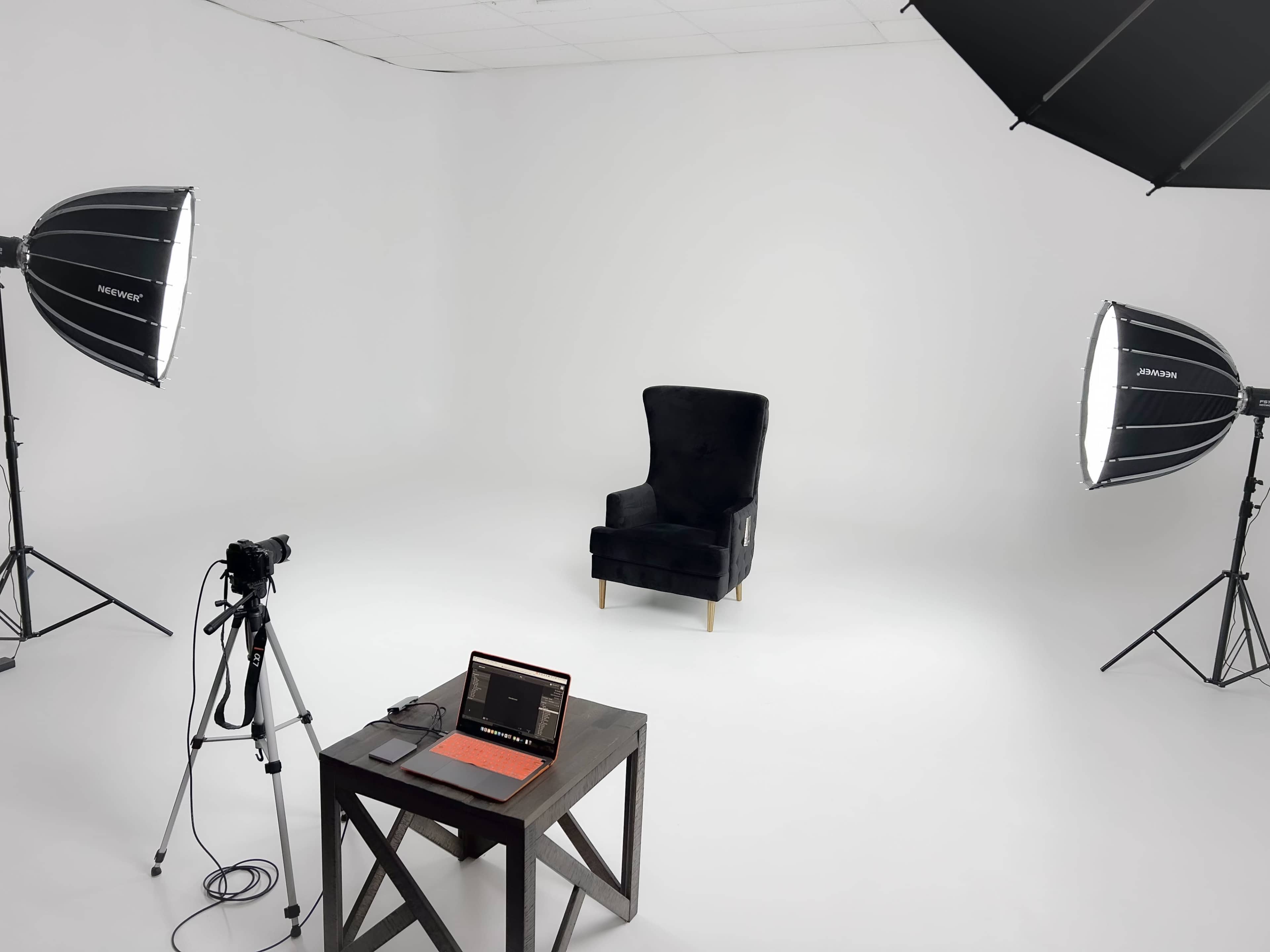 A minimalist studio setup with a black velvet chair, a camera on a tripod, and a laptop on a wooden table, illuminated by two softbox lights.