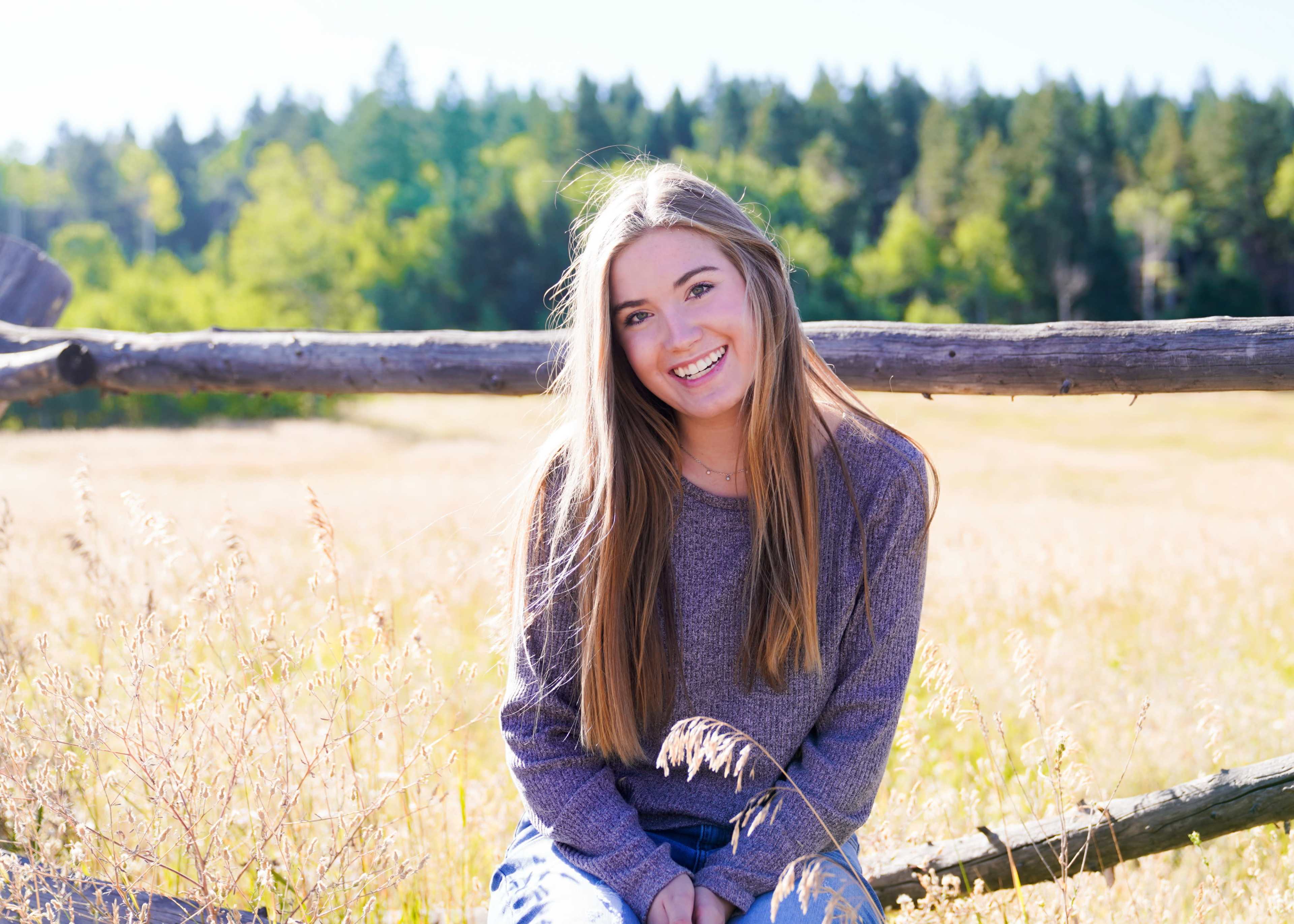 A young woman with long hair sits on a wooden fence in a grassy field surrounded by trees.