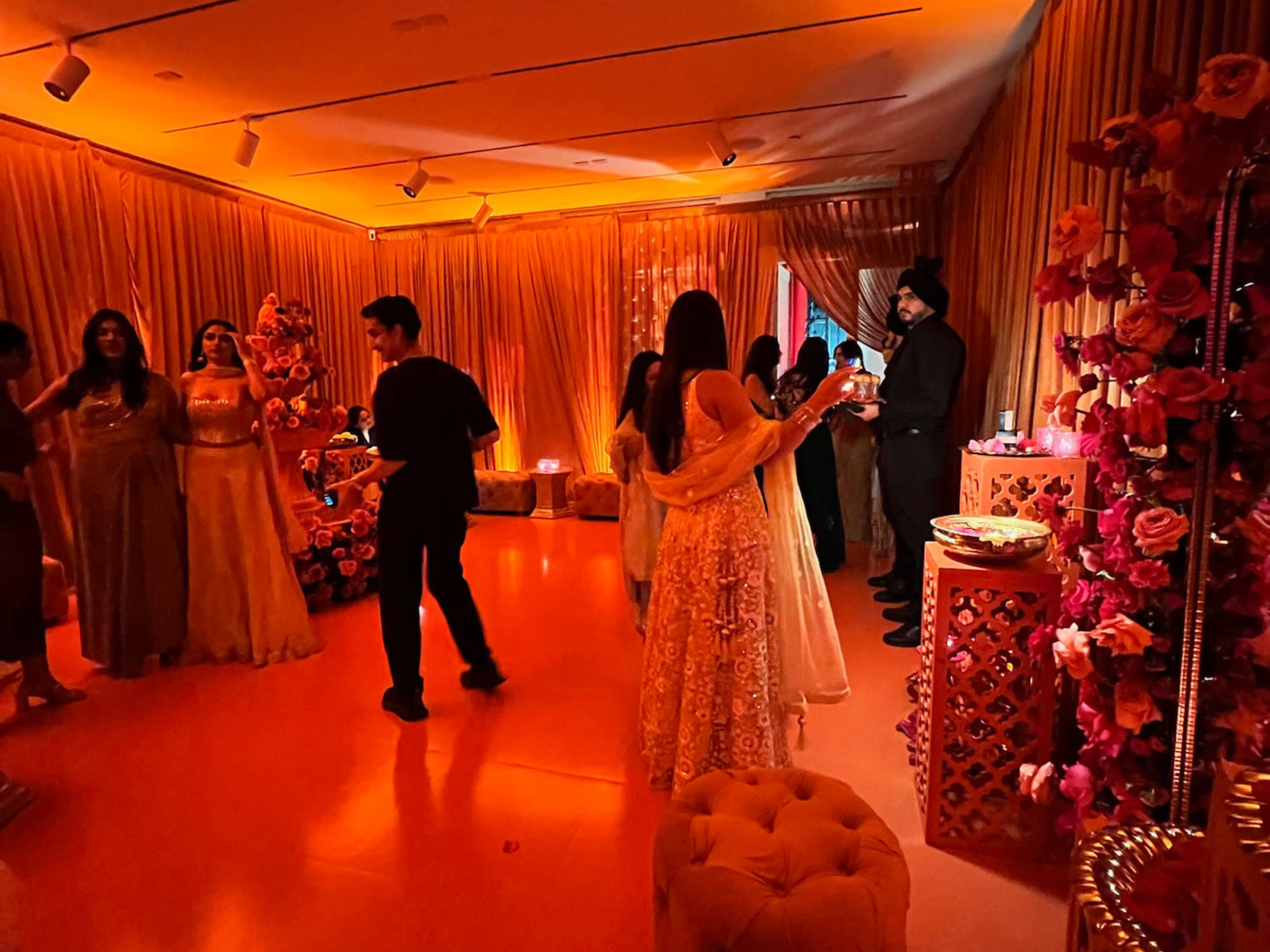 A group of elegantly dressed people socializes in a warmly lit, floral-decorated room with orange-toned lighting.