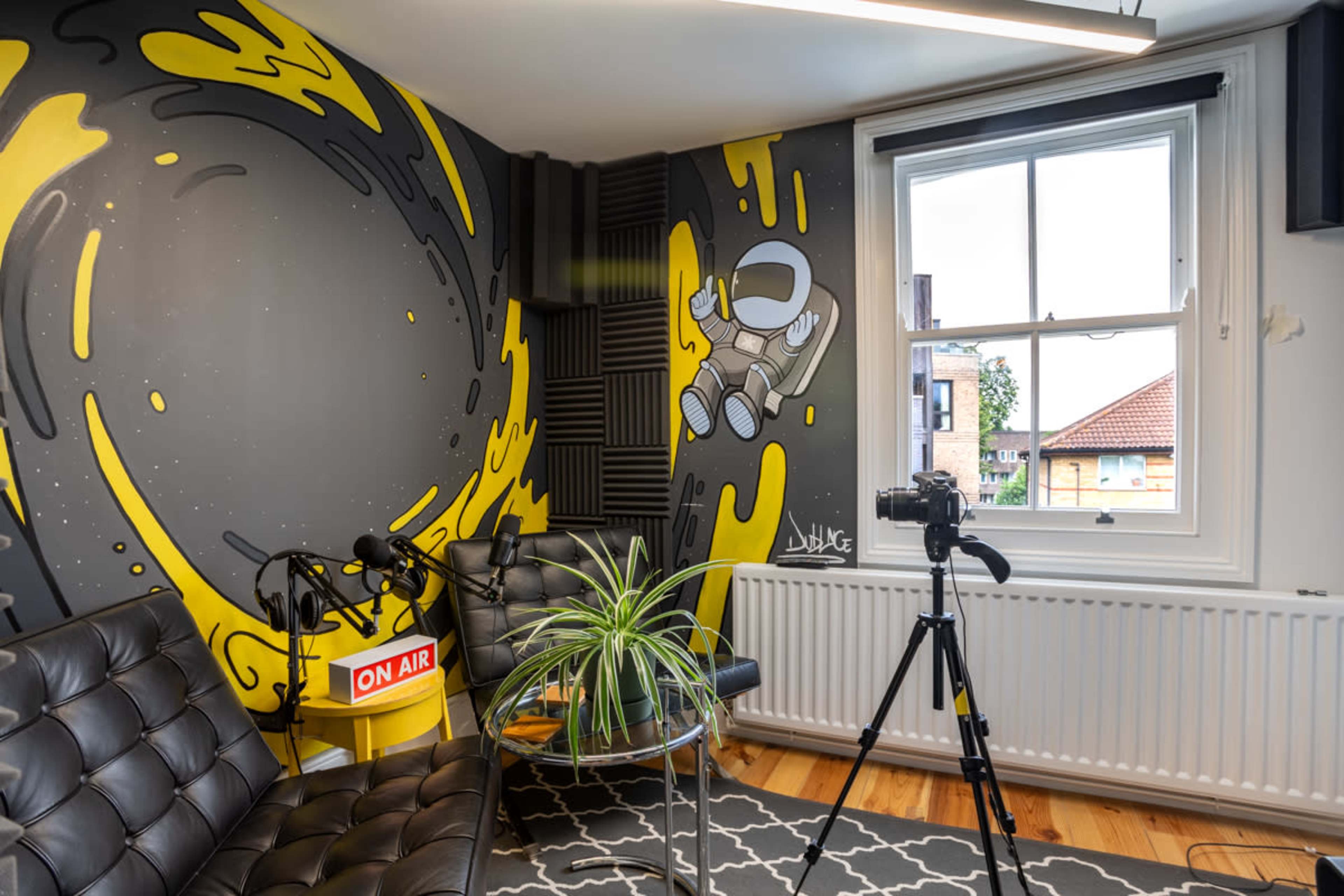 A recording studio featuring a wall with a yellow and gray mural, black seating, and equipment including microphones and a camera.