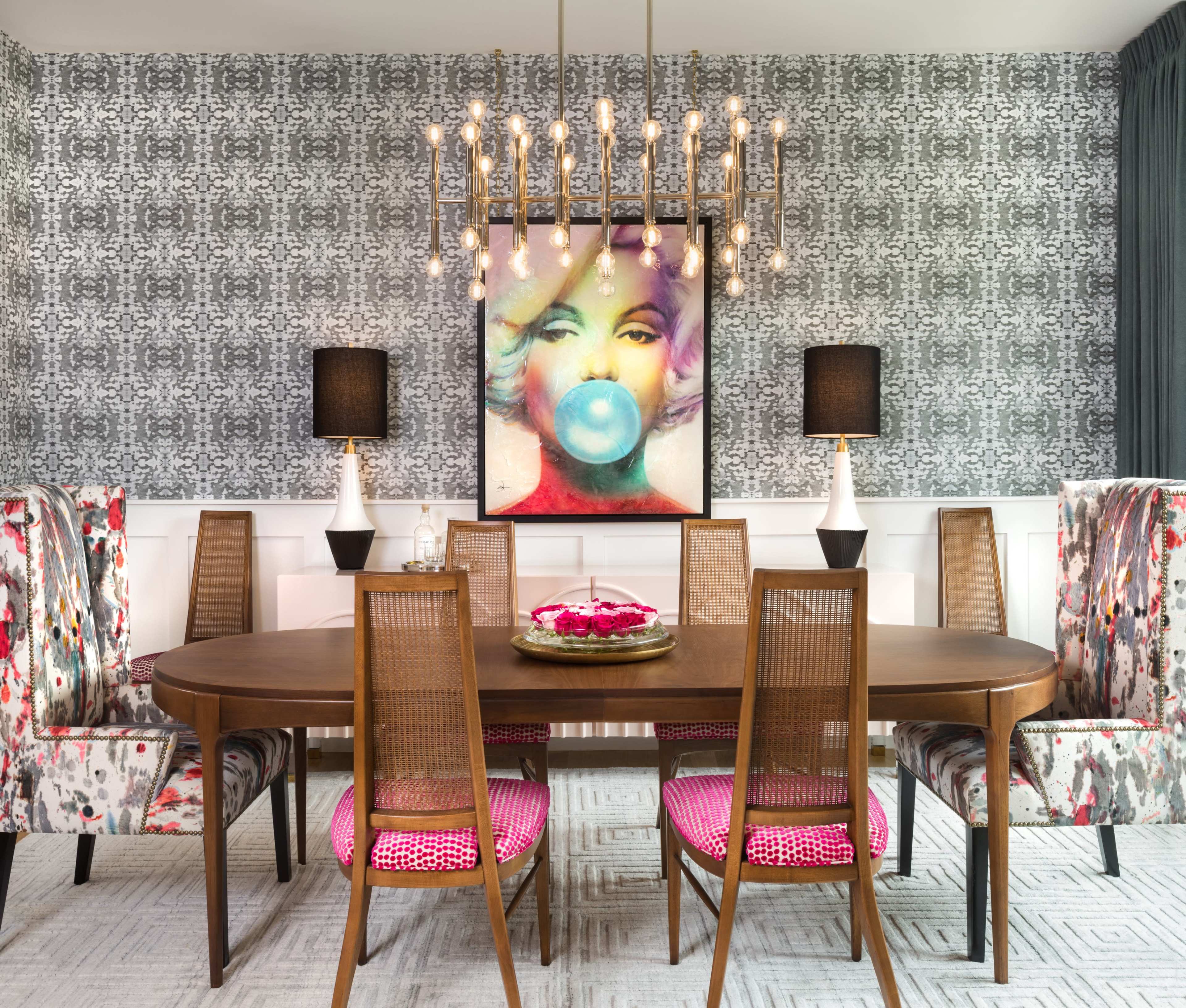 The image features a modern dining room with a circular wooden table surrounded by various chairs, a large artwork of a woman blowing a bubble, and a contemporary chandelier above.