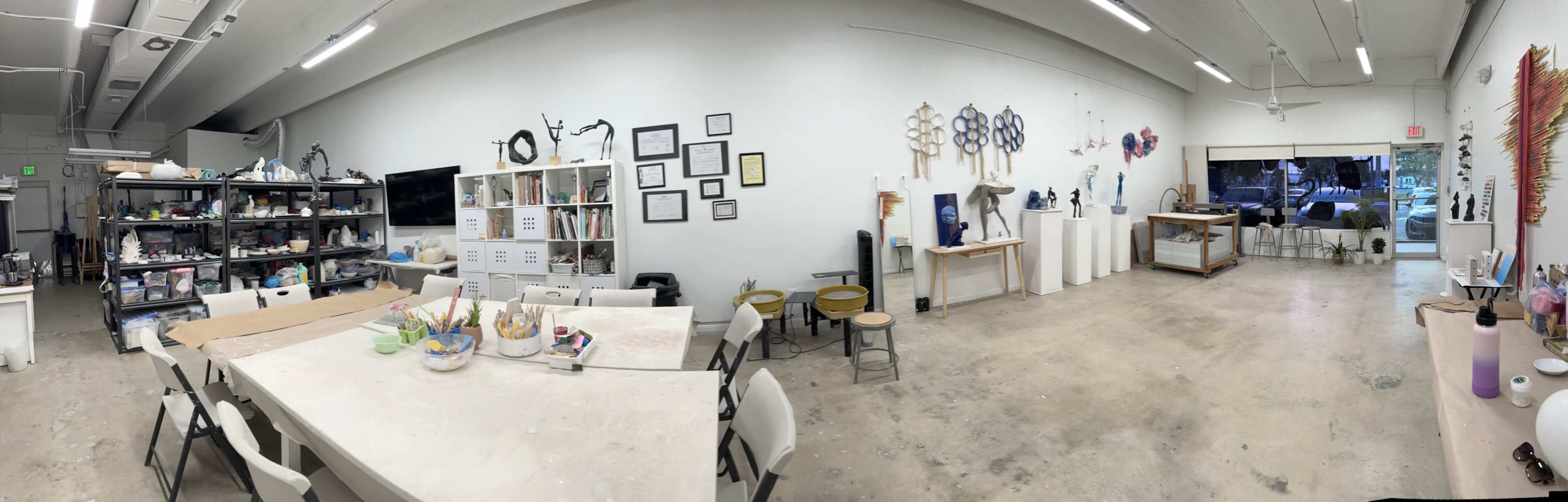 The image shows an open art studio featuring tables, chairs, shelves filled with art supplies, and various artworks displayed on the walls.