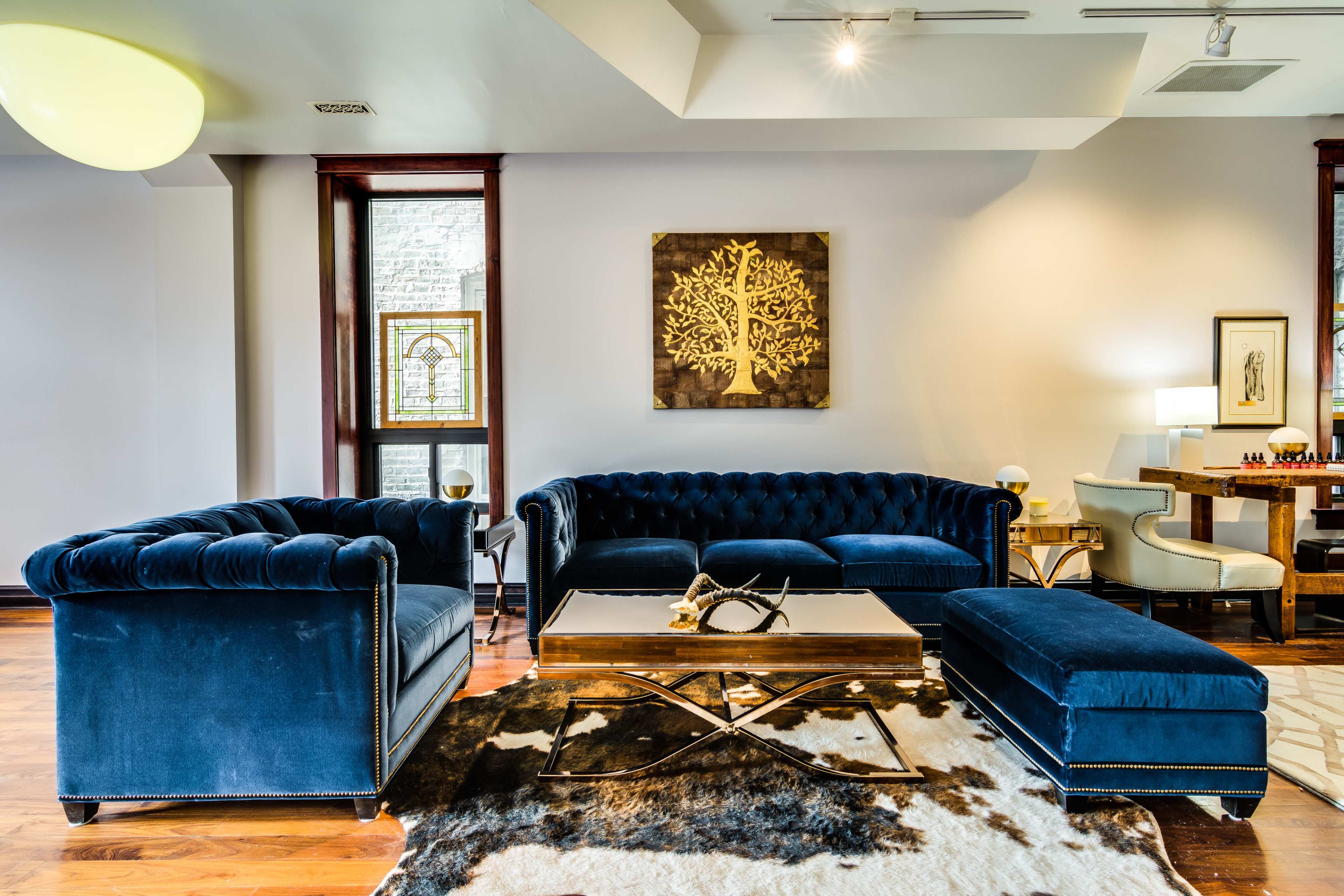 A modern living room features blue velvet furniture arranged around a coffee table with a geometric design, complemented by a light-colored artwork on the wall and a wooden floor.