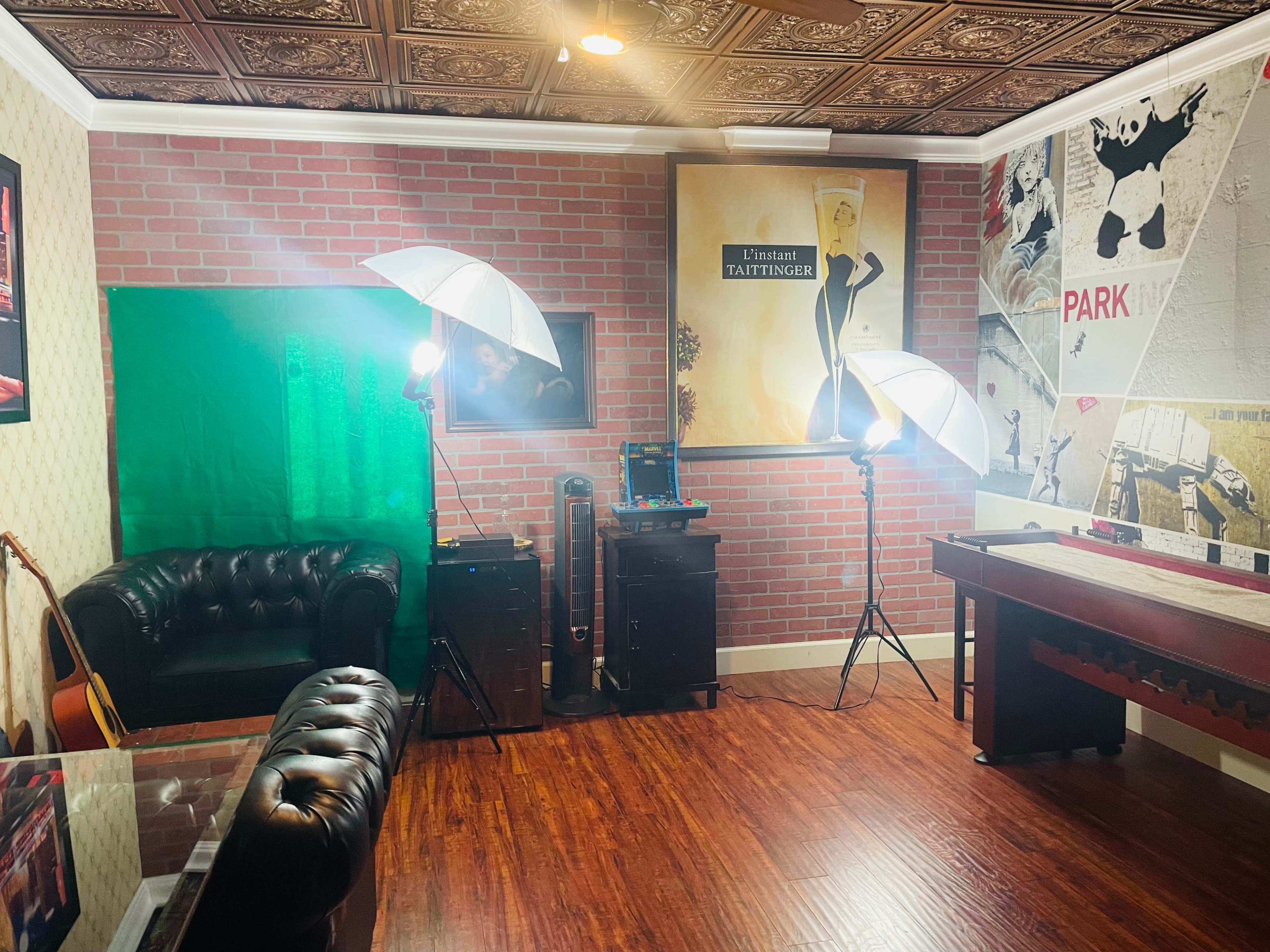 The image depicts a room with a brick wall, a green screen setup, photography lights, a leather couch, and posters on the walls.
