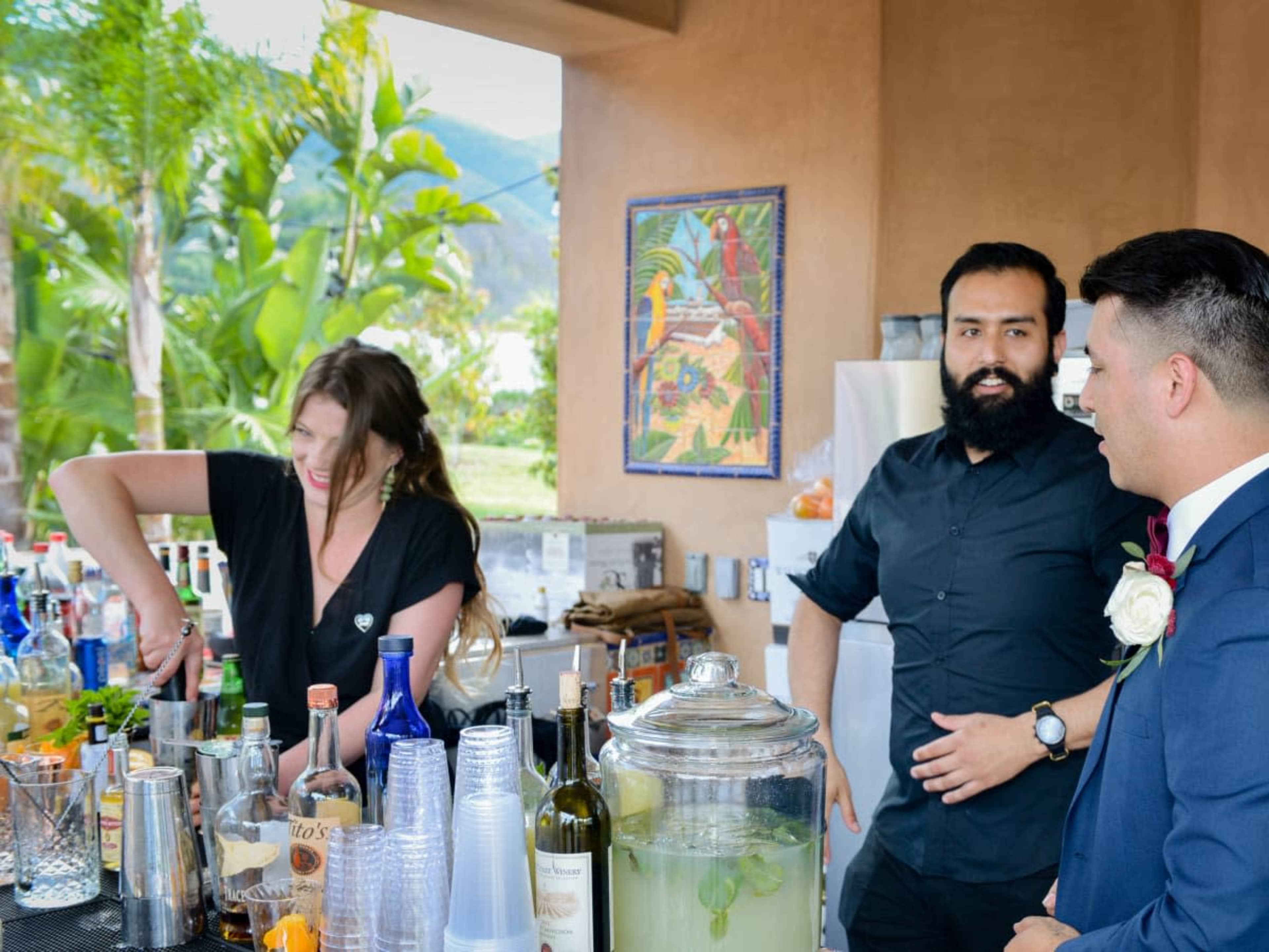 A bartender prepares a drink while a man in formal attire converses with a colleague at an outdoor bar surrounded by tropical plants.