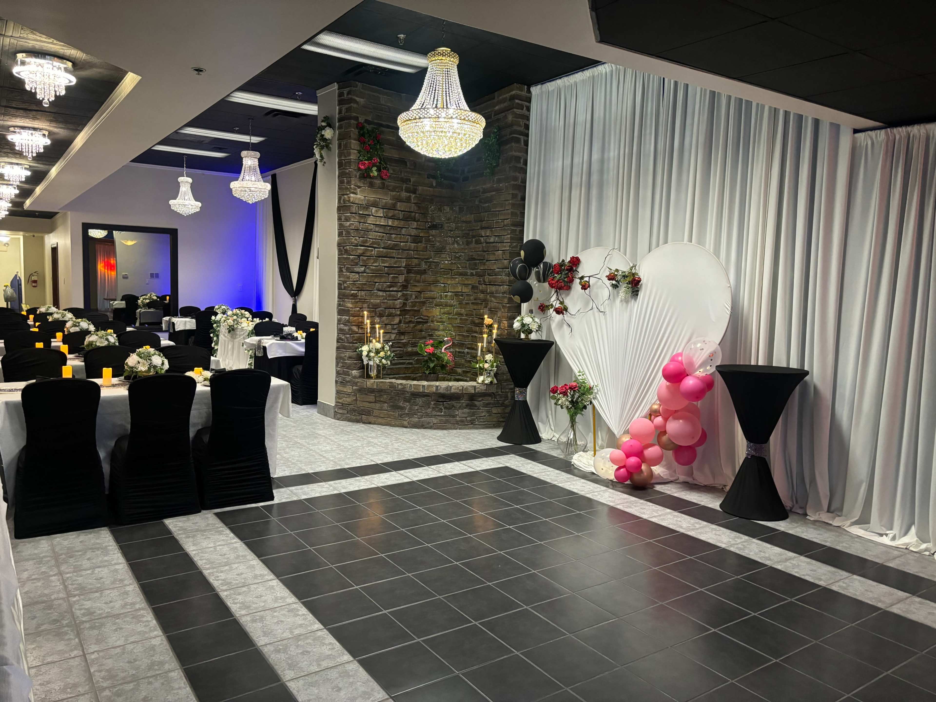 The image shows a decorated event space with black and white tables, a floral backdrop with balloons, and chandeliers hanging from the ceiling.