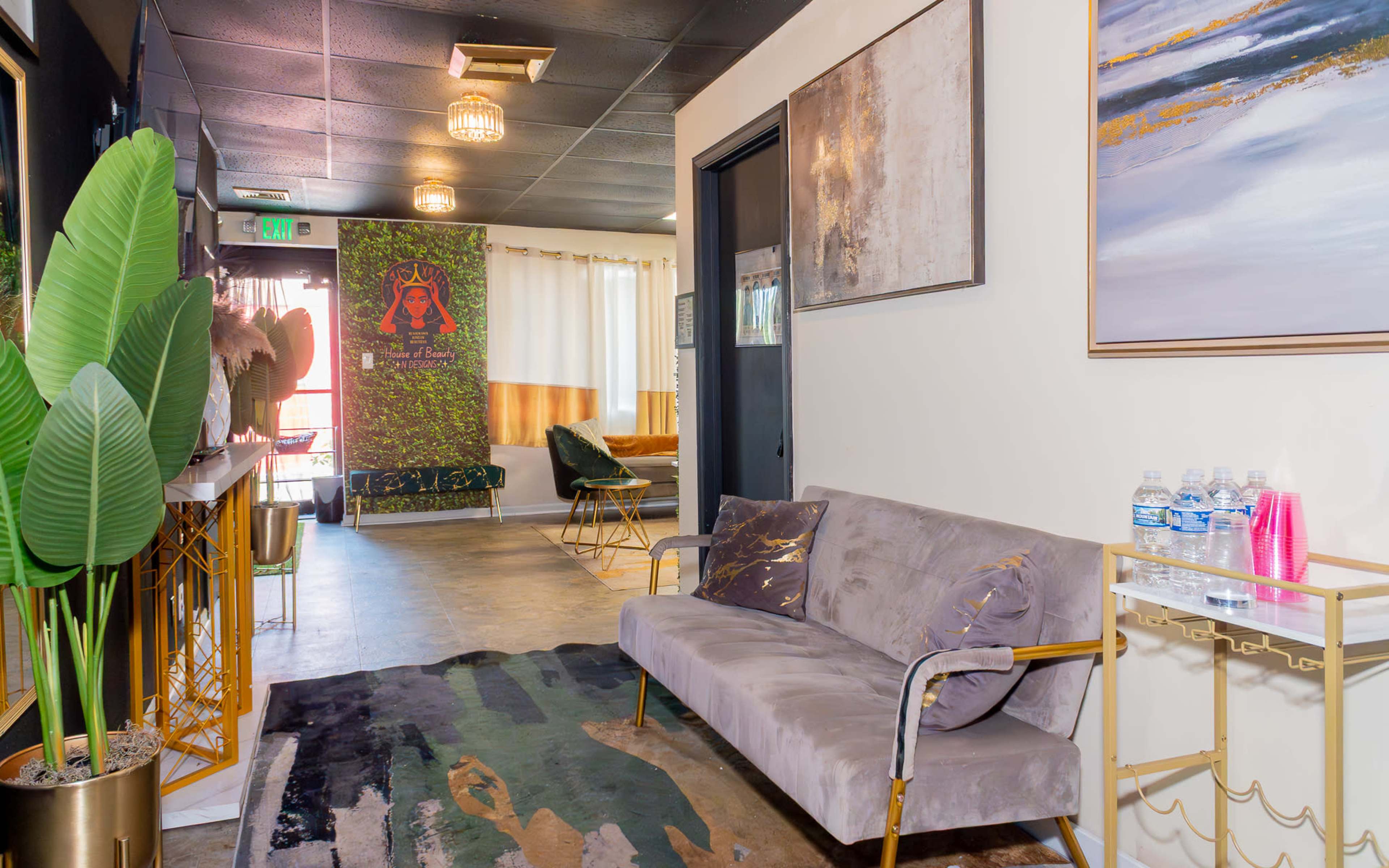 The image shows a modern hallway featuring a plush gray sofa, large potted plants, decorative artwork, and water bottles on a side table.