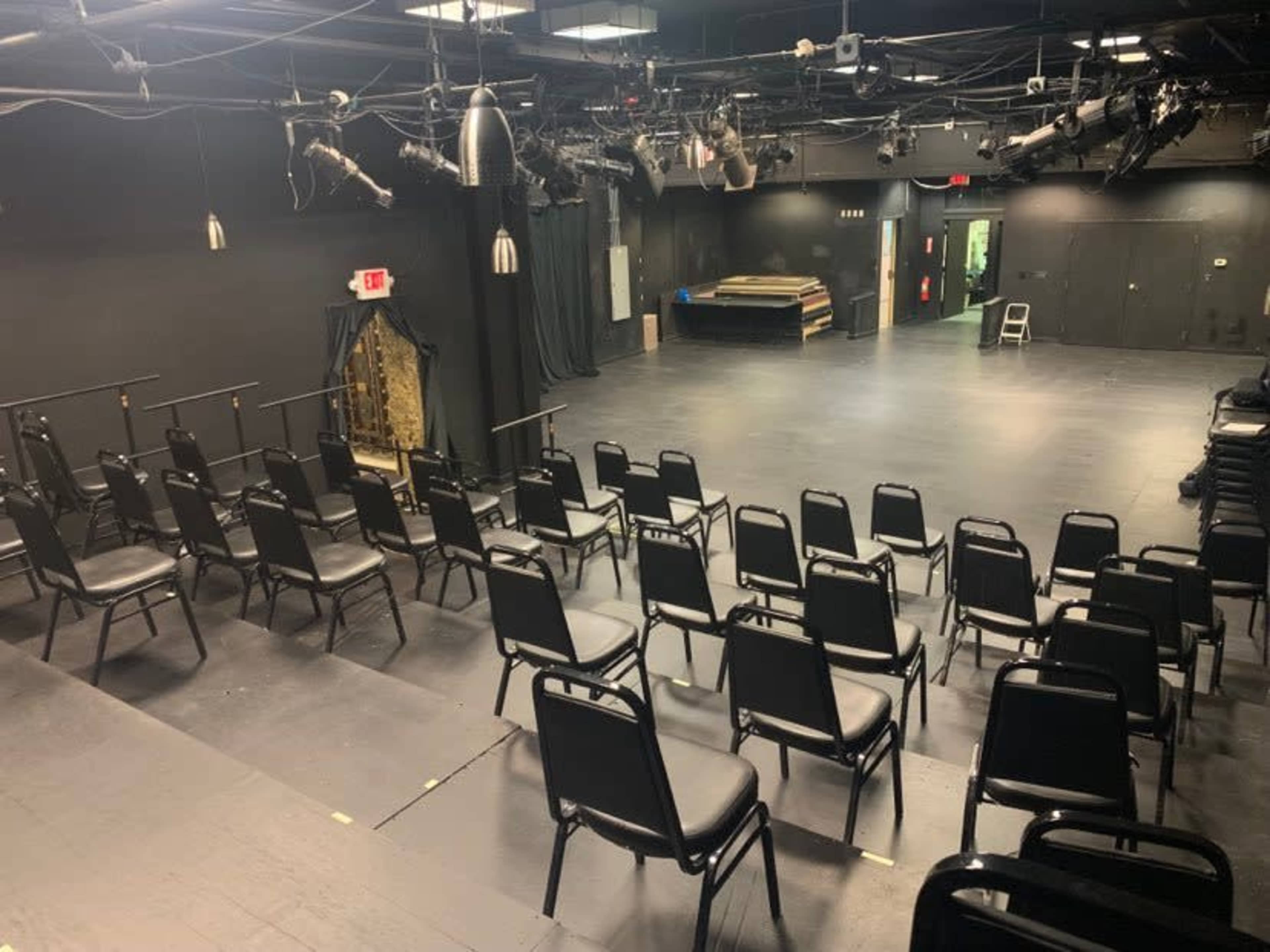 The image shows a small black box theater with rows of black chairs facing a performance space illuminated by overhead stage lights.