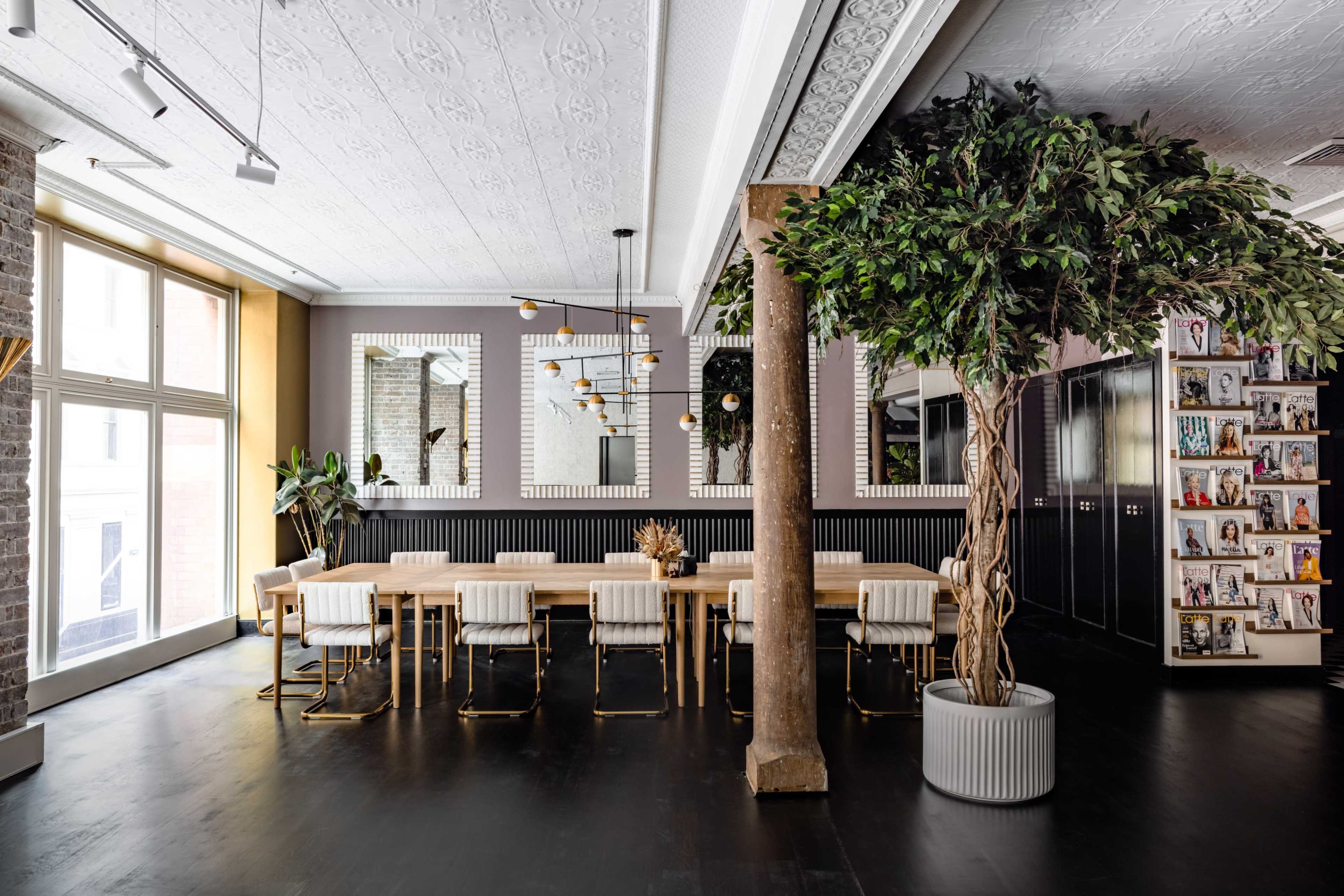 A modern dining area features a long wooden table surrounded by white chairs, with large windows and a tall potted plant enhancing the space.