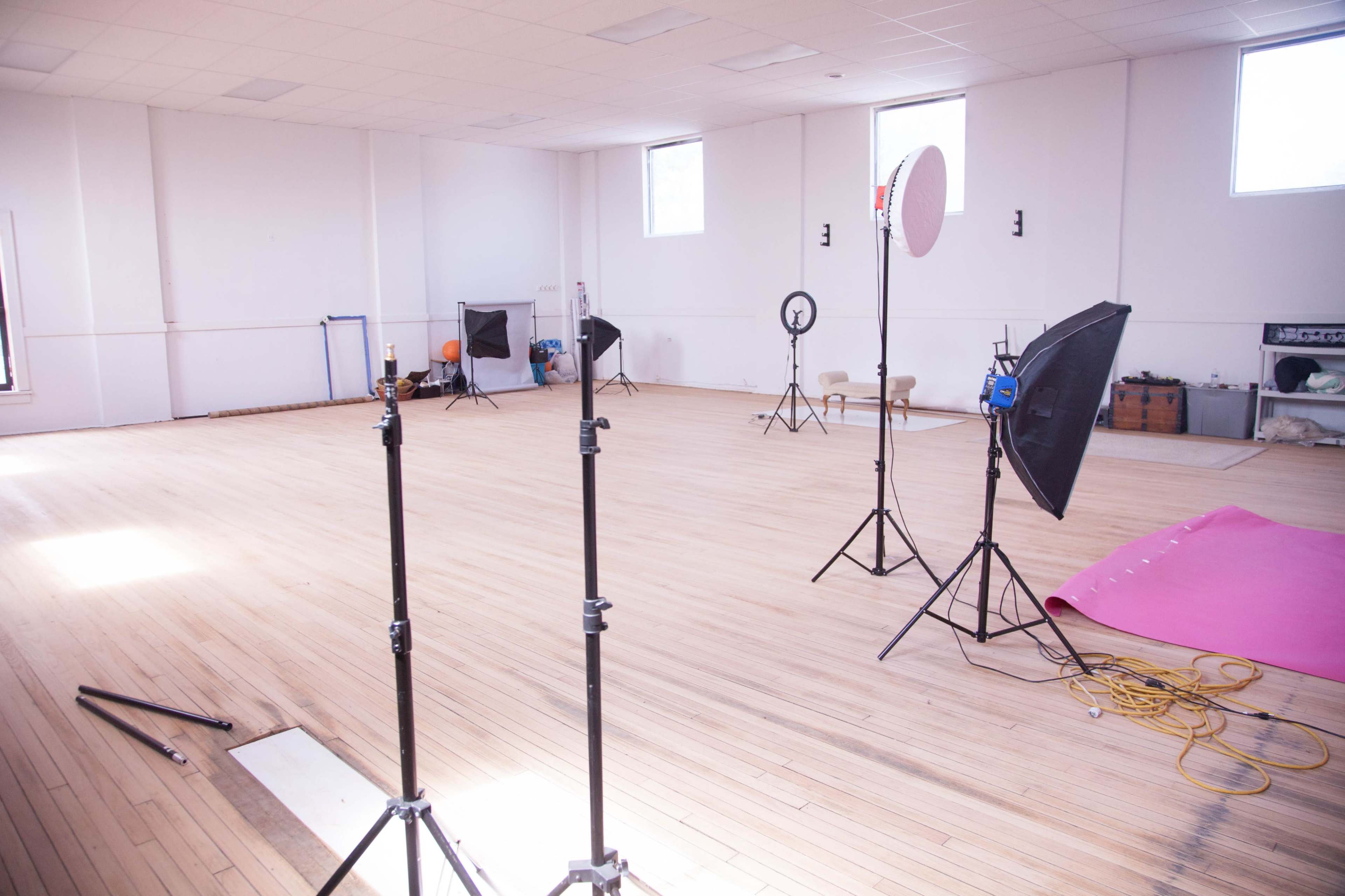 A spacious, empty photography studio with various lighting equipment and backdrops set up around a wooden floor.