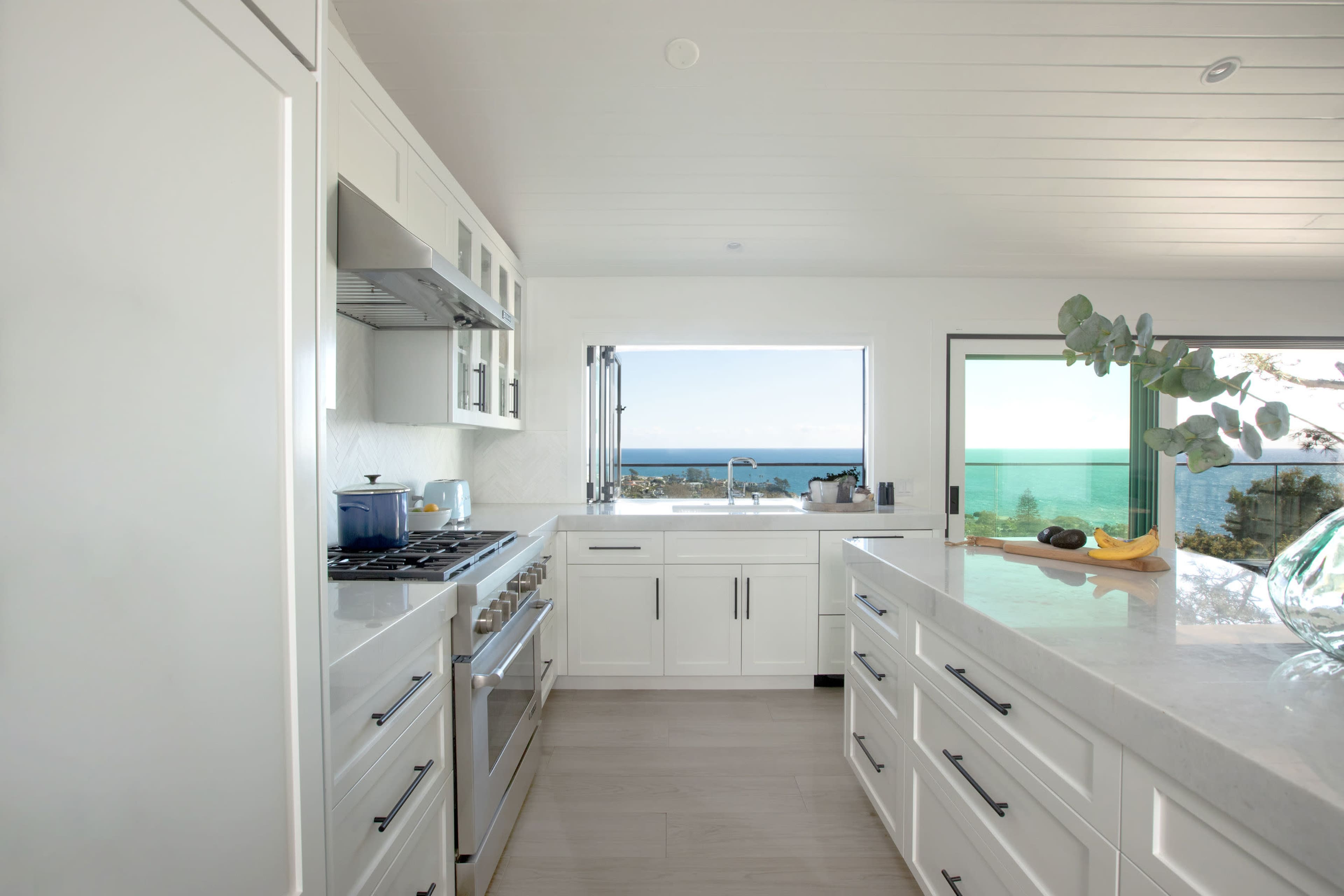 The image shows a modern kitchen with white cabinetry, a large window showcasing an ocean view, and stainless steel appliances.