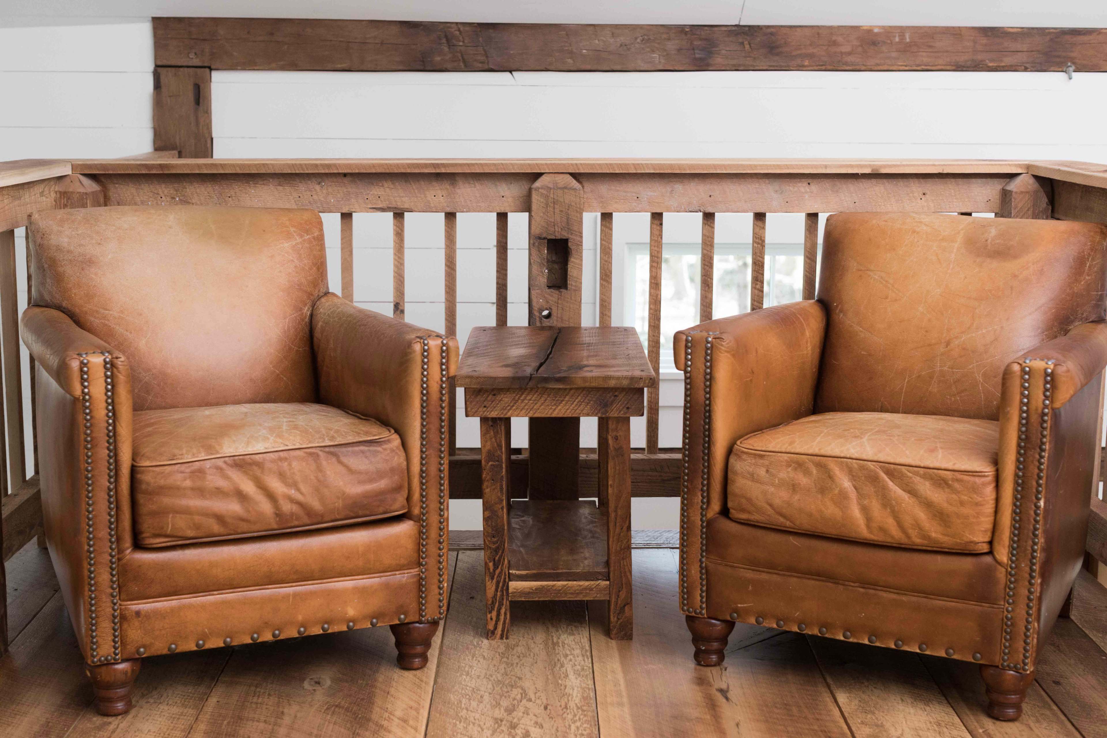 Two leather armchairs are positioned on a wooden floor beside a small wooden side table, set against a backdrop of wooden railings.