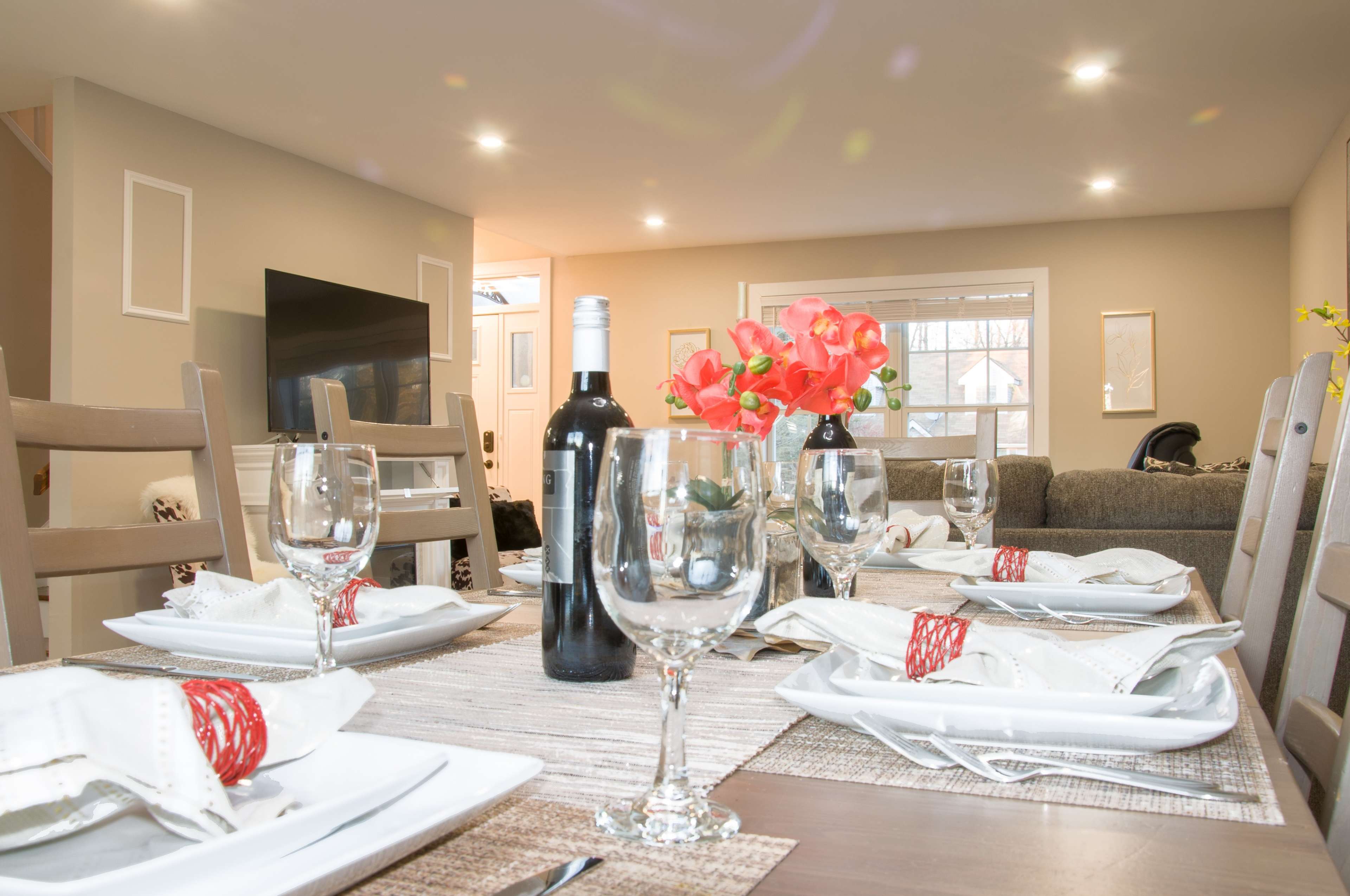 A dining table is set with wine bottles, glasses, and neatly arranged plates, while a cozy living area with a television is visible in the background.