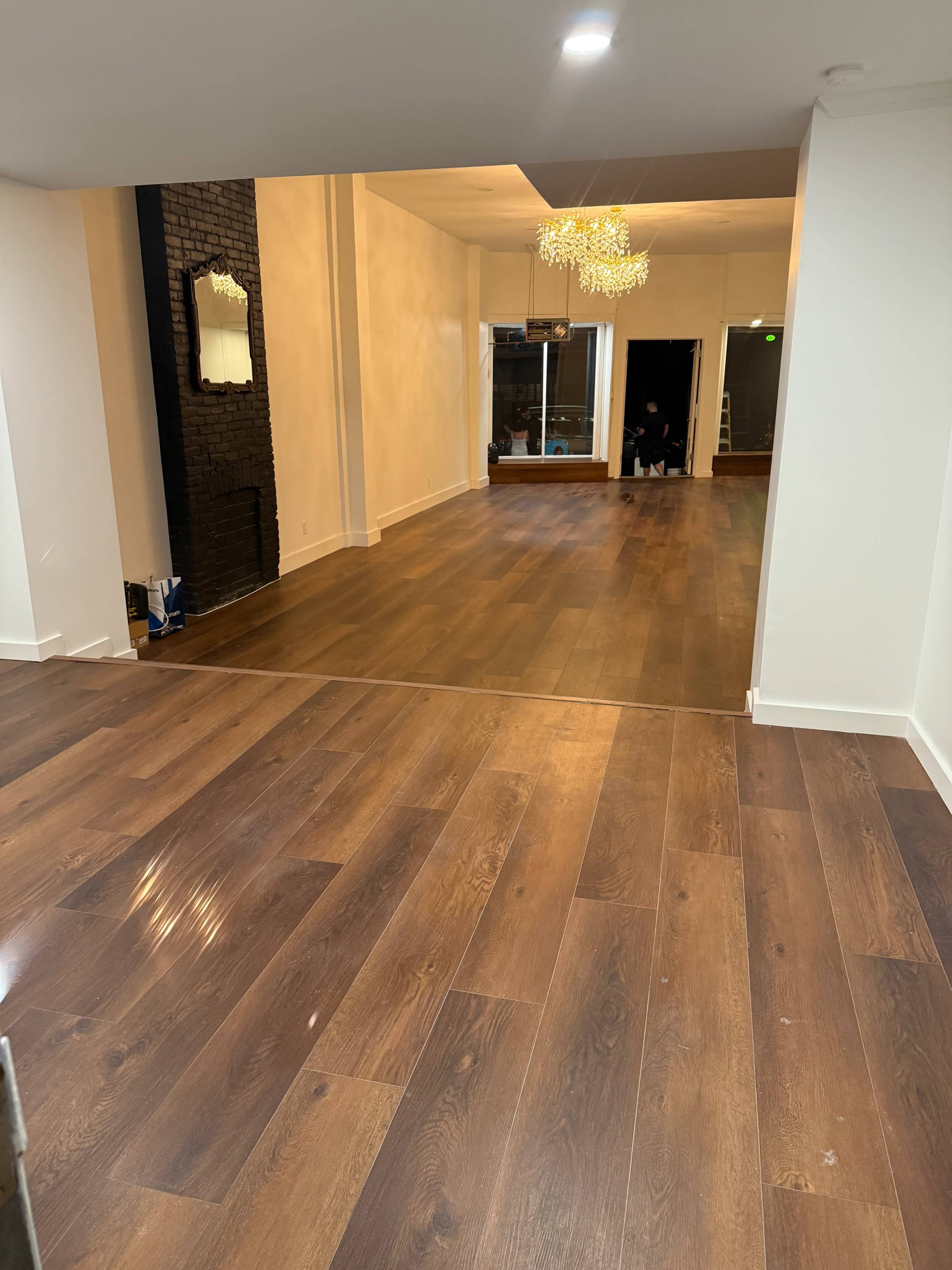 The image shows a spacious interior with hardwood flooring, featuring a large mirror on one wall and a chandelier hanging from the ceiling.