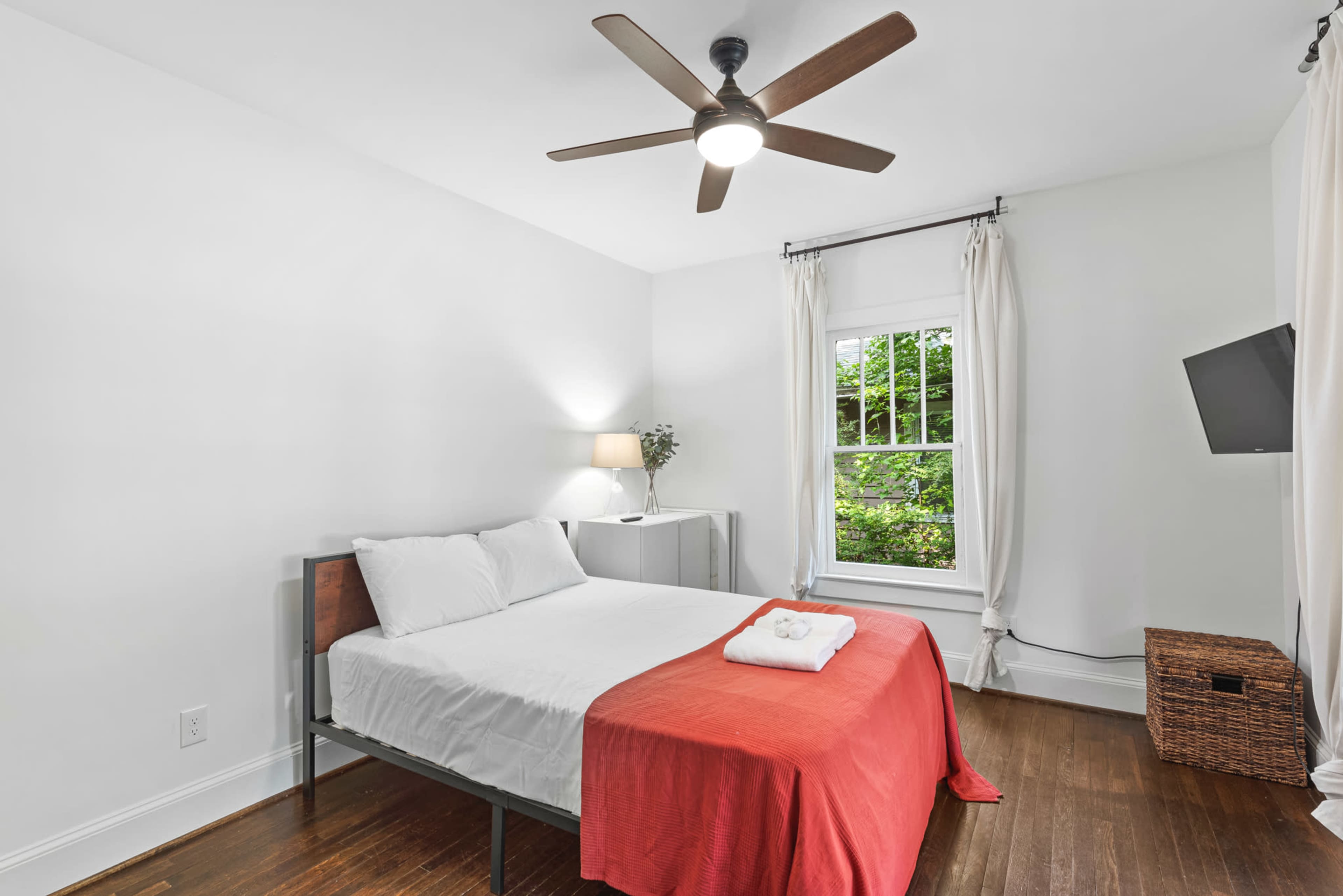 A tidy bedroom features a double bed with white linens and a red throw, a ceiling fan, a nightstand with a lamp, and a window draped with white curtains.