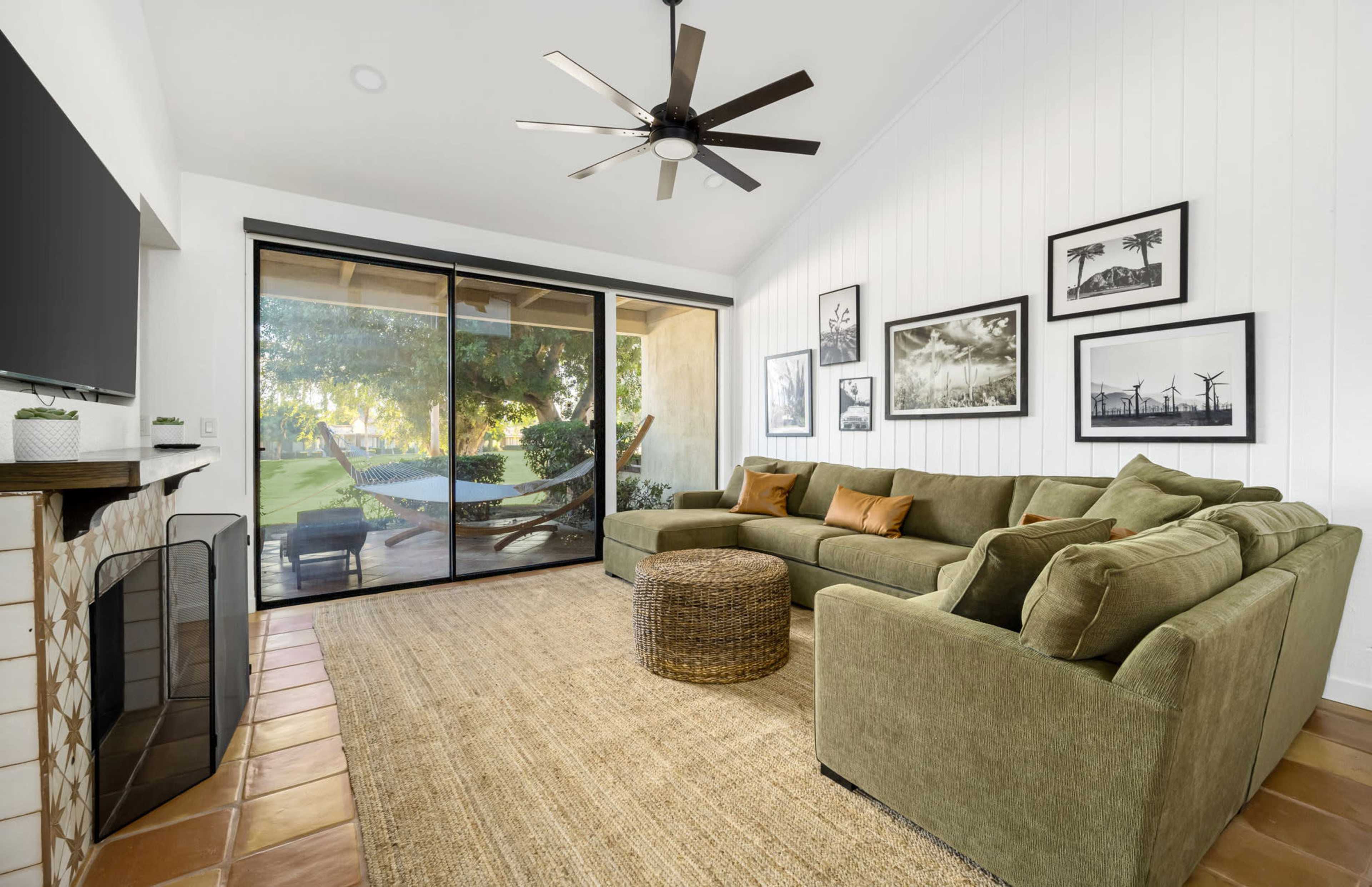 The living room features a green sectional sofa, a round woven coffee table, and large sliding glass doors that open to a patio with a view of a garden.