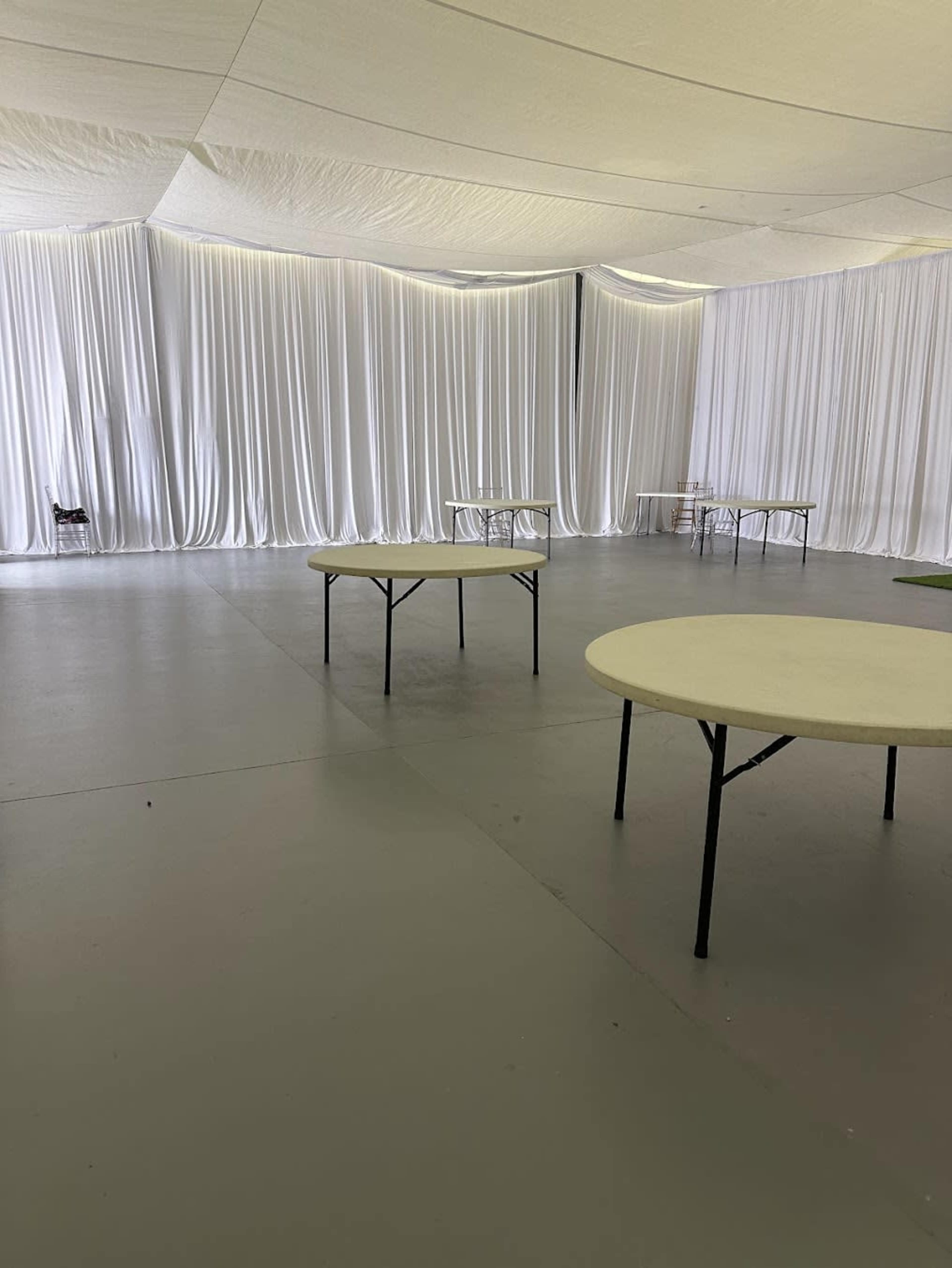 The image shows a large, empty indoor space with white draped curtains and several round tables arranged throughout.