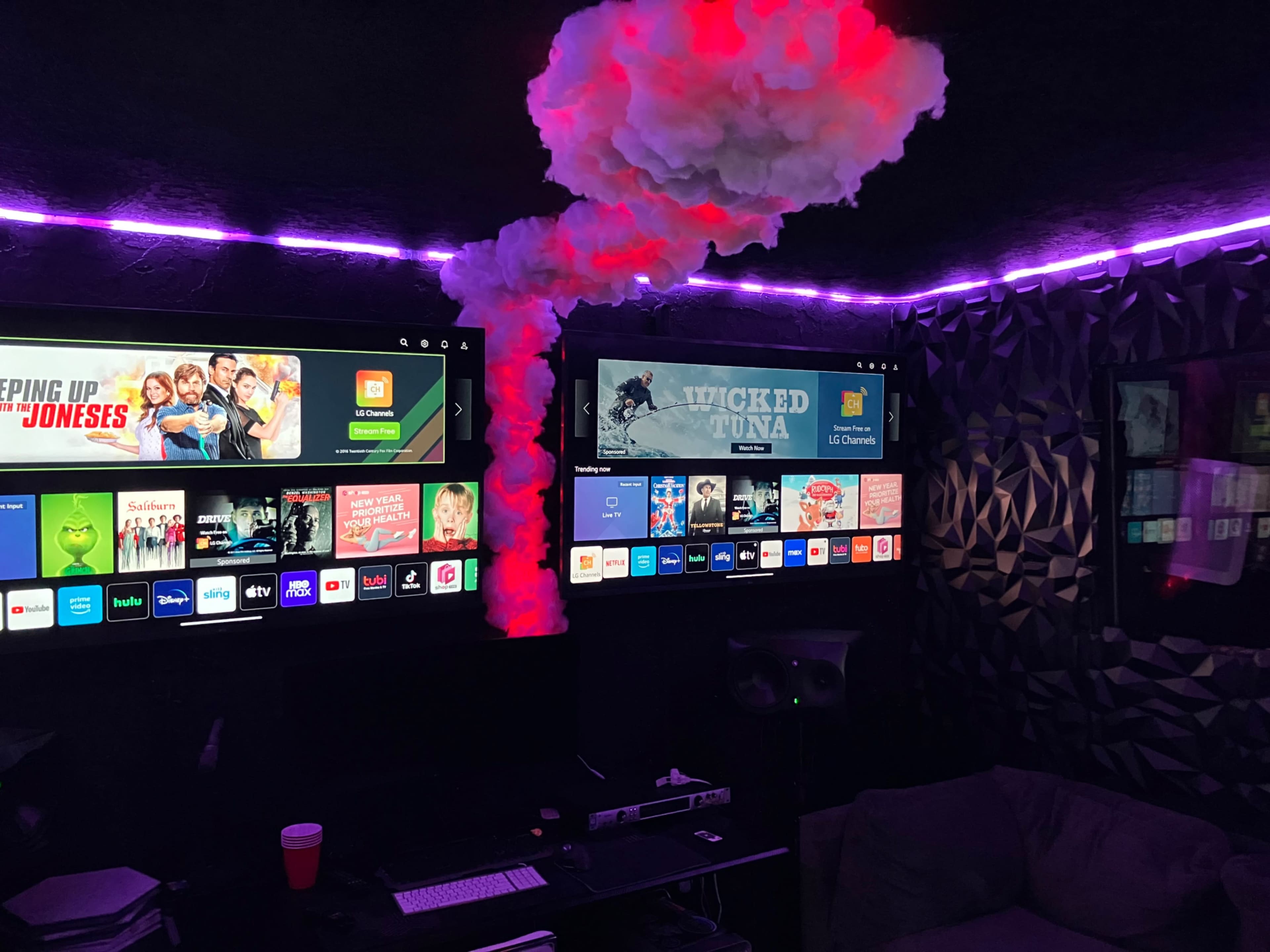 The image shows a media room with two large flat-screen TVs displaying different content under purple ambient lighting, surrounded by a cloud-like installation on the ceiling.