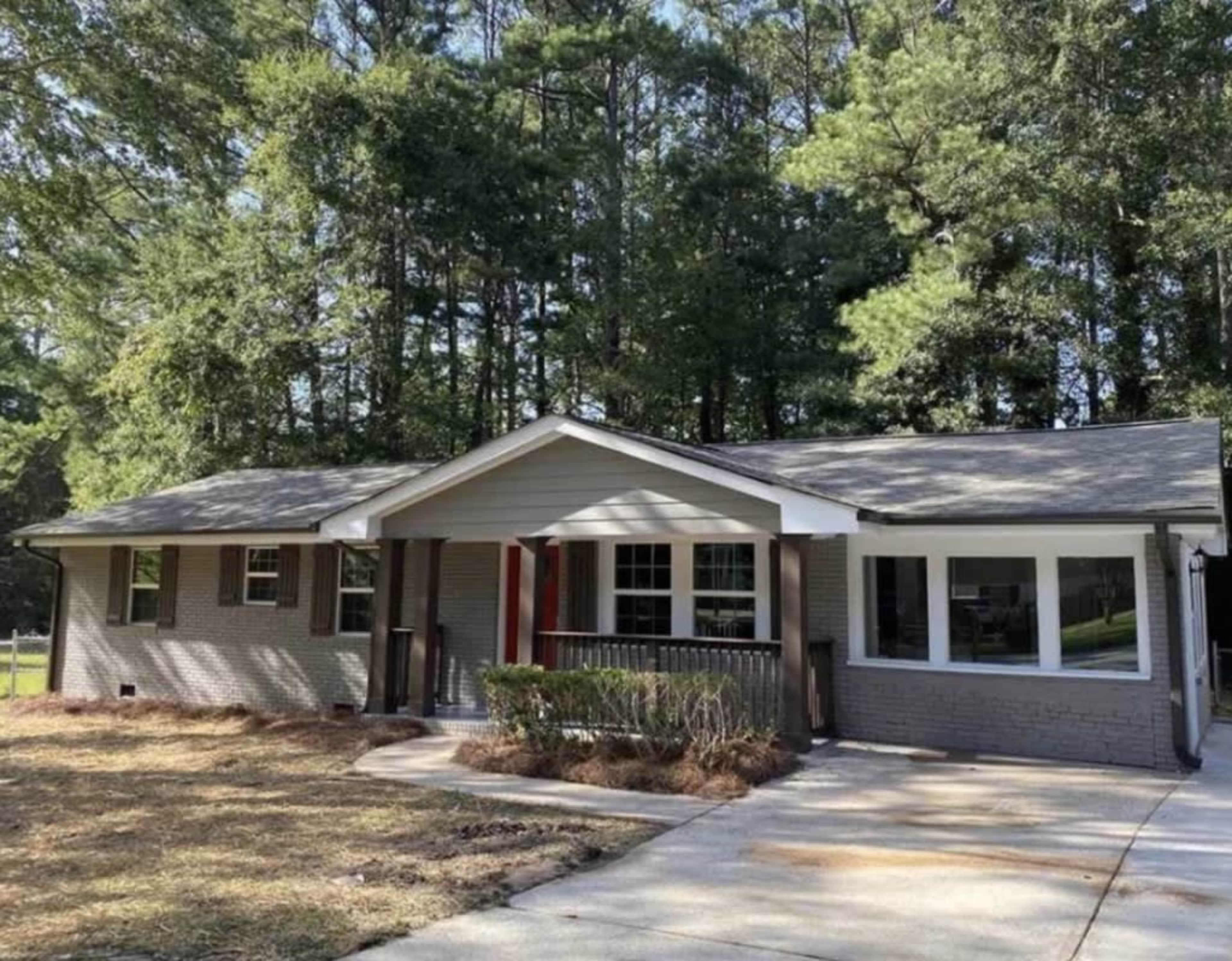 A single-story gray brick house with a front porch sits amidst tall trees, featuring multiple windows and a concrete driveway.