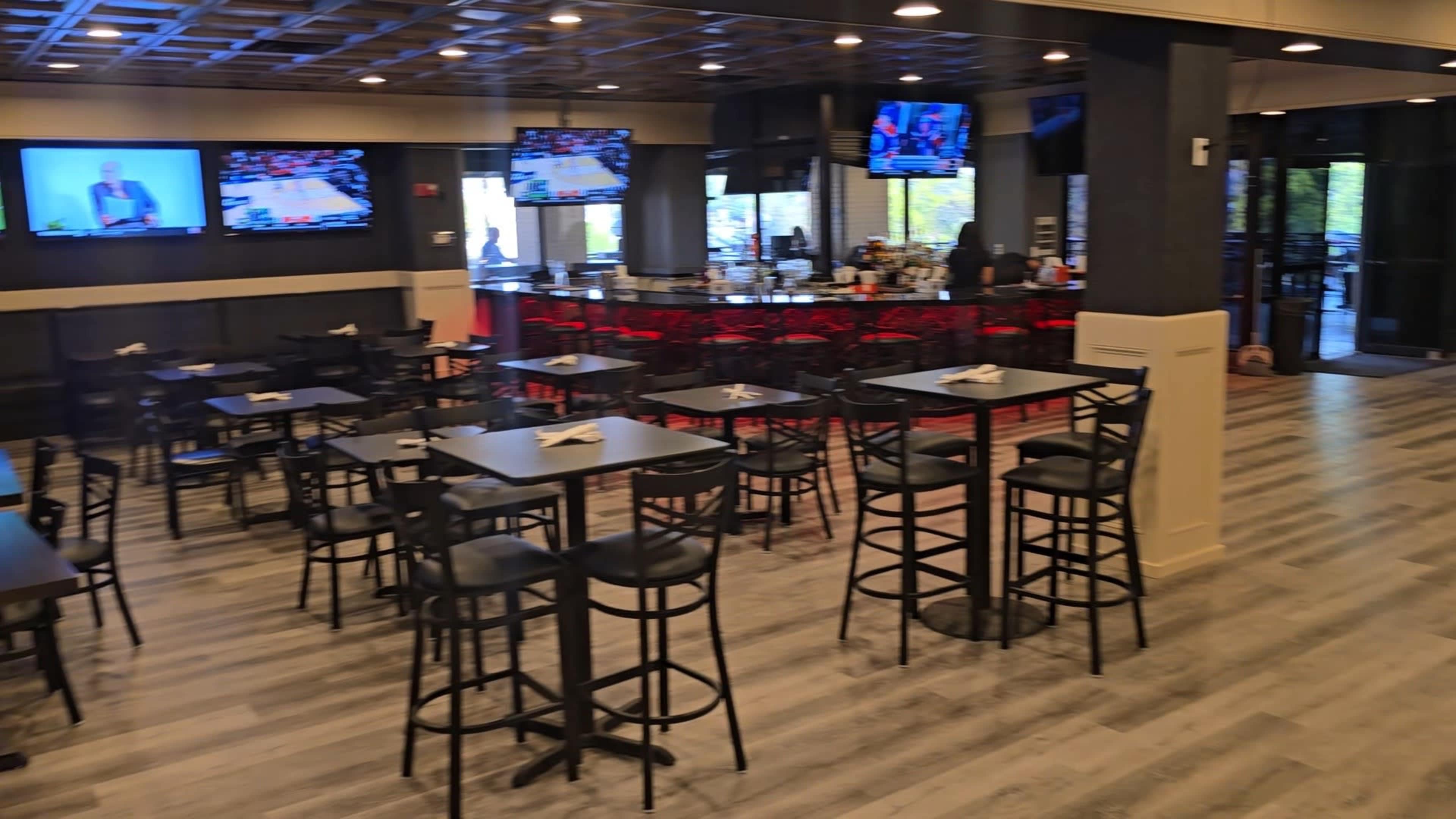 The image shows a modern bar and dining area with several tables and high chairs, where multiple televisions display sports events.