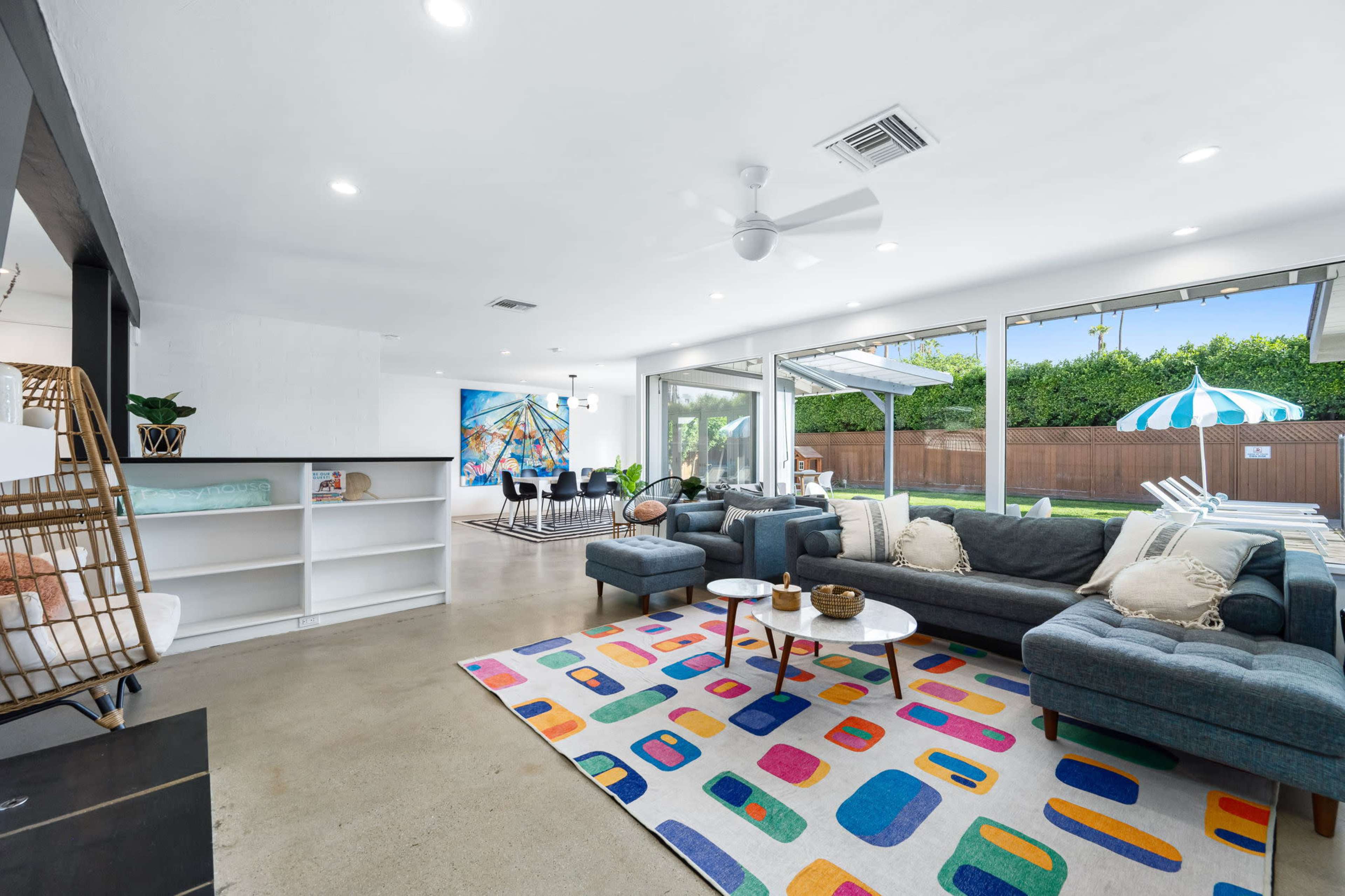A spacious living area features a gray sectional sofa, vibrant area rug, and large windows overlooking a patio with a swimming pool.