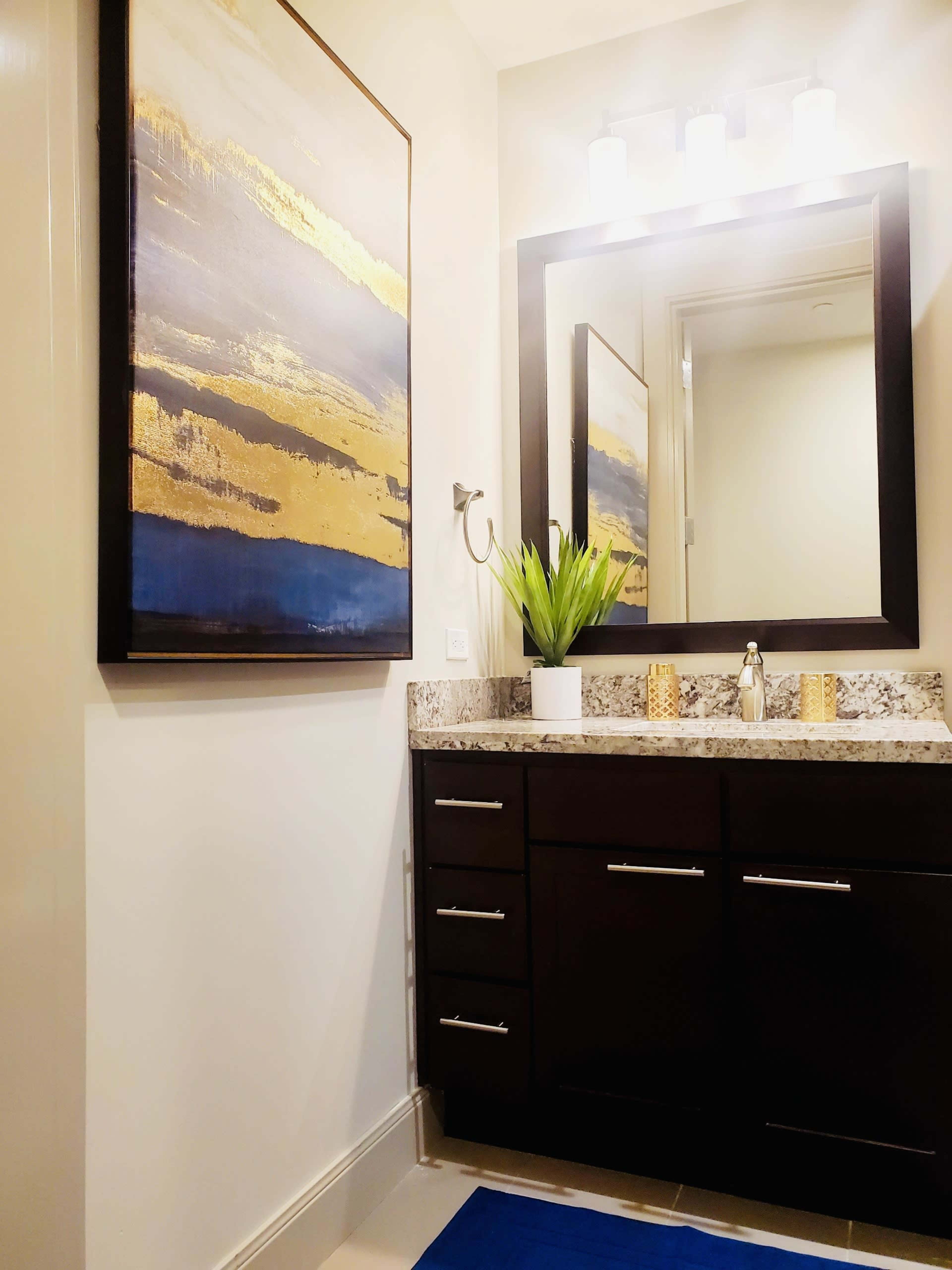 A modern bathroom with a dark wood vanity, a large mirror, and a piece of artwork featuring gold and blue hues on the wall.