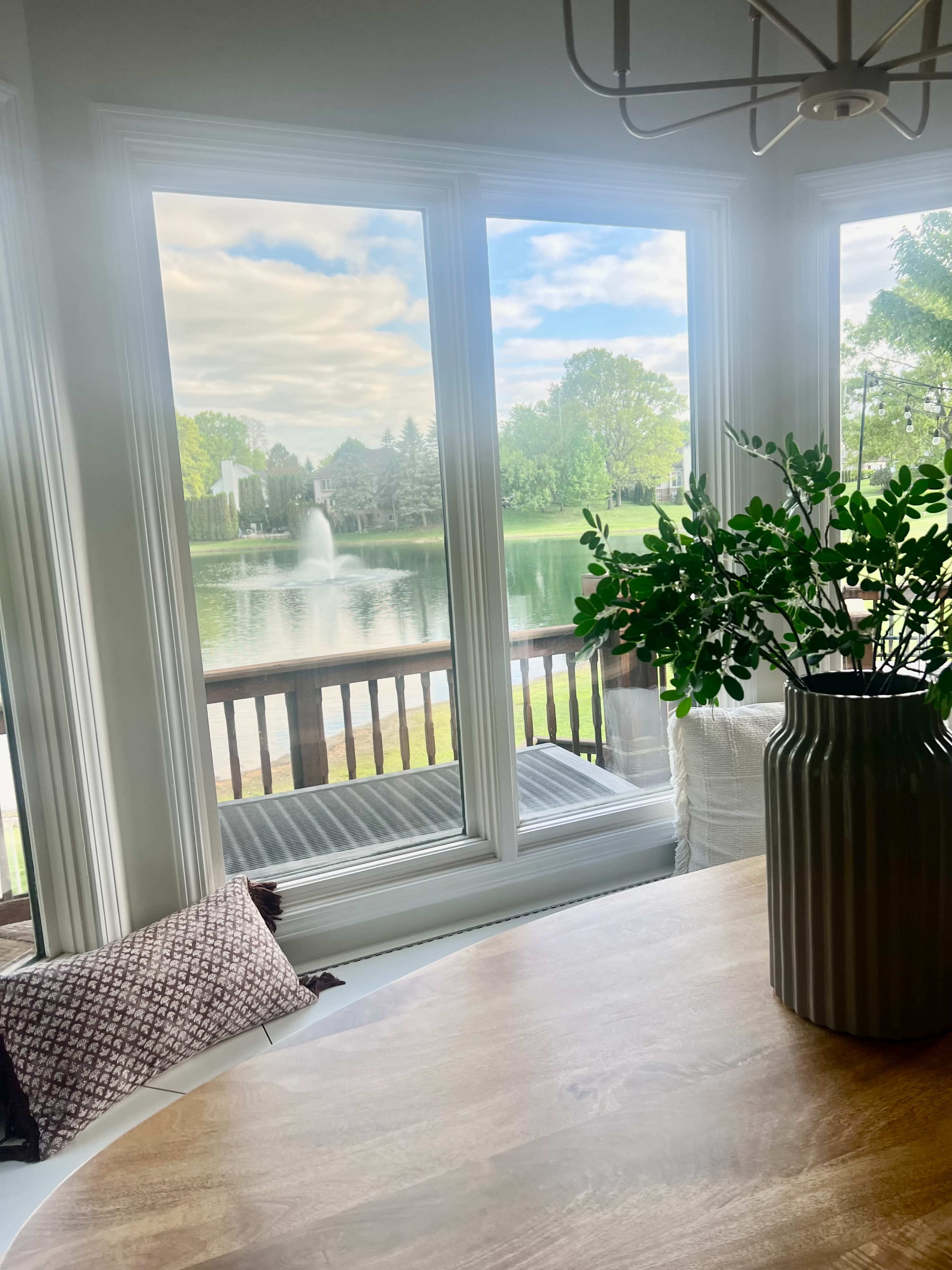 A view from a dining area features large windows overlooking a serene lake with a fountain and green trees in the background.