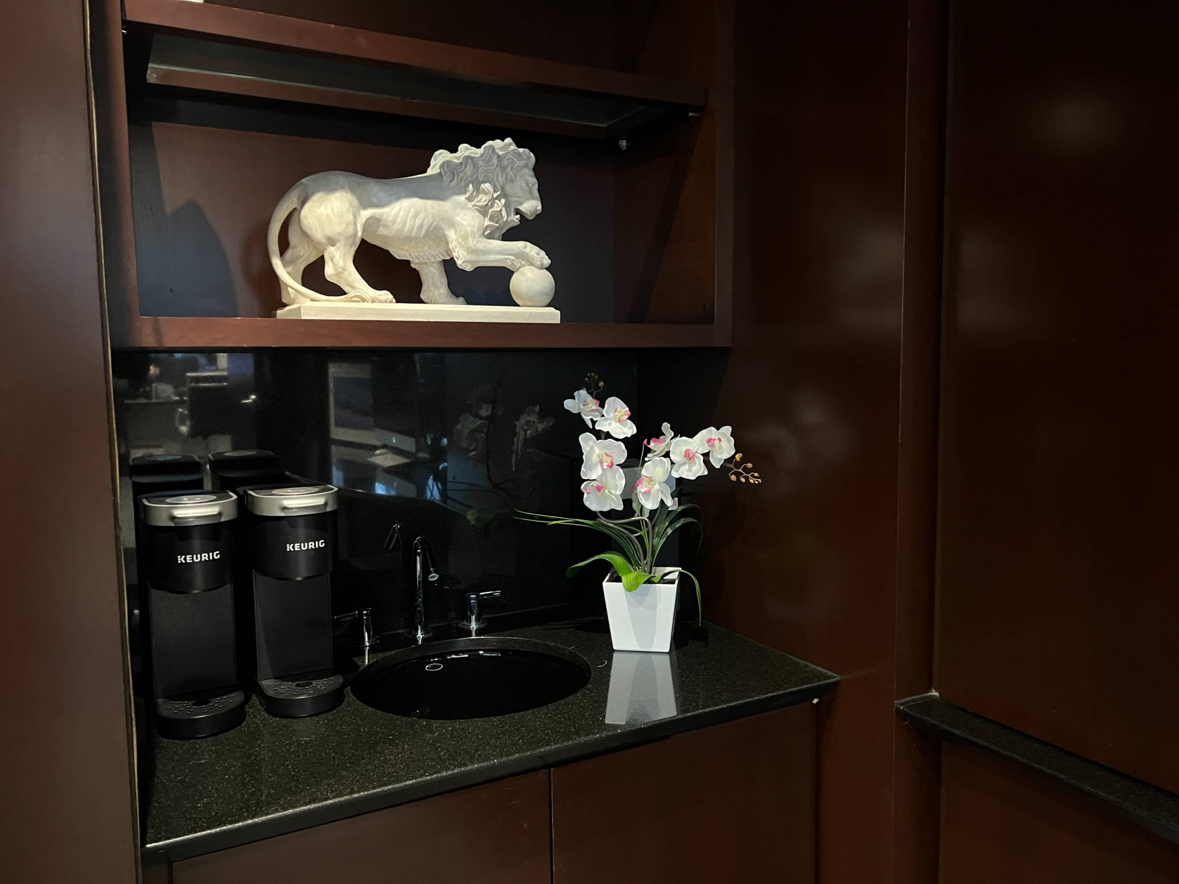 A dark wooden cabinet featuring a lion sculpture, two coffee machines, a small sink, and a potted orchid plant.