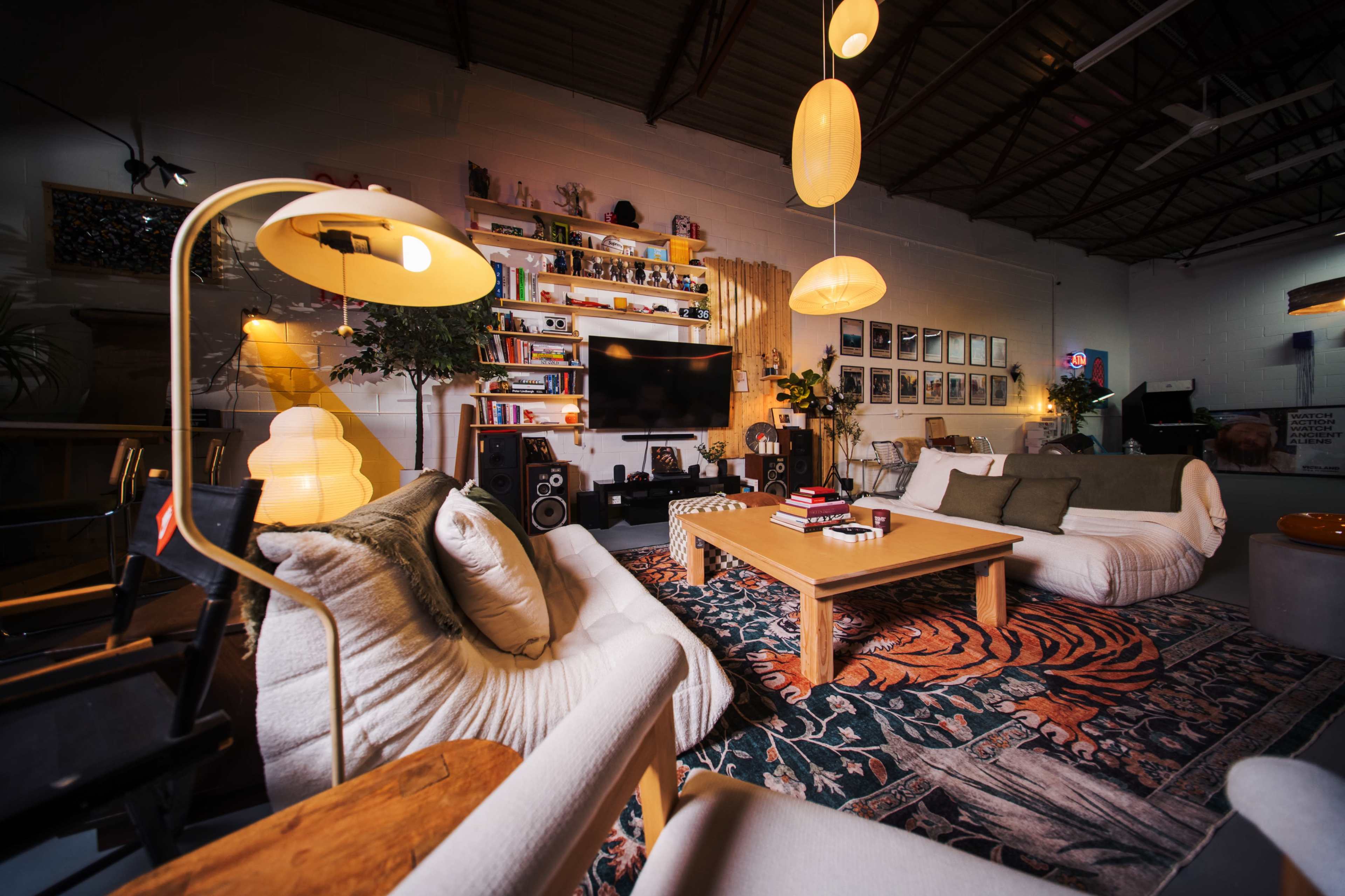 The image shows a modern living room with two couches, a coffee table in the center, a large television mounted on the wall, and various decorative items and plants arranged throughout the space.
