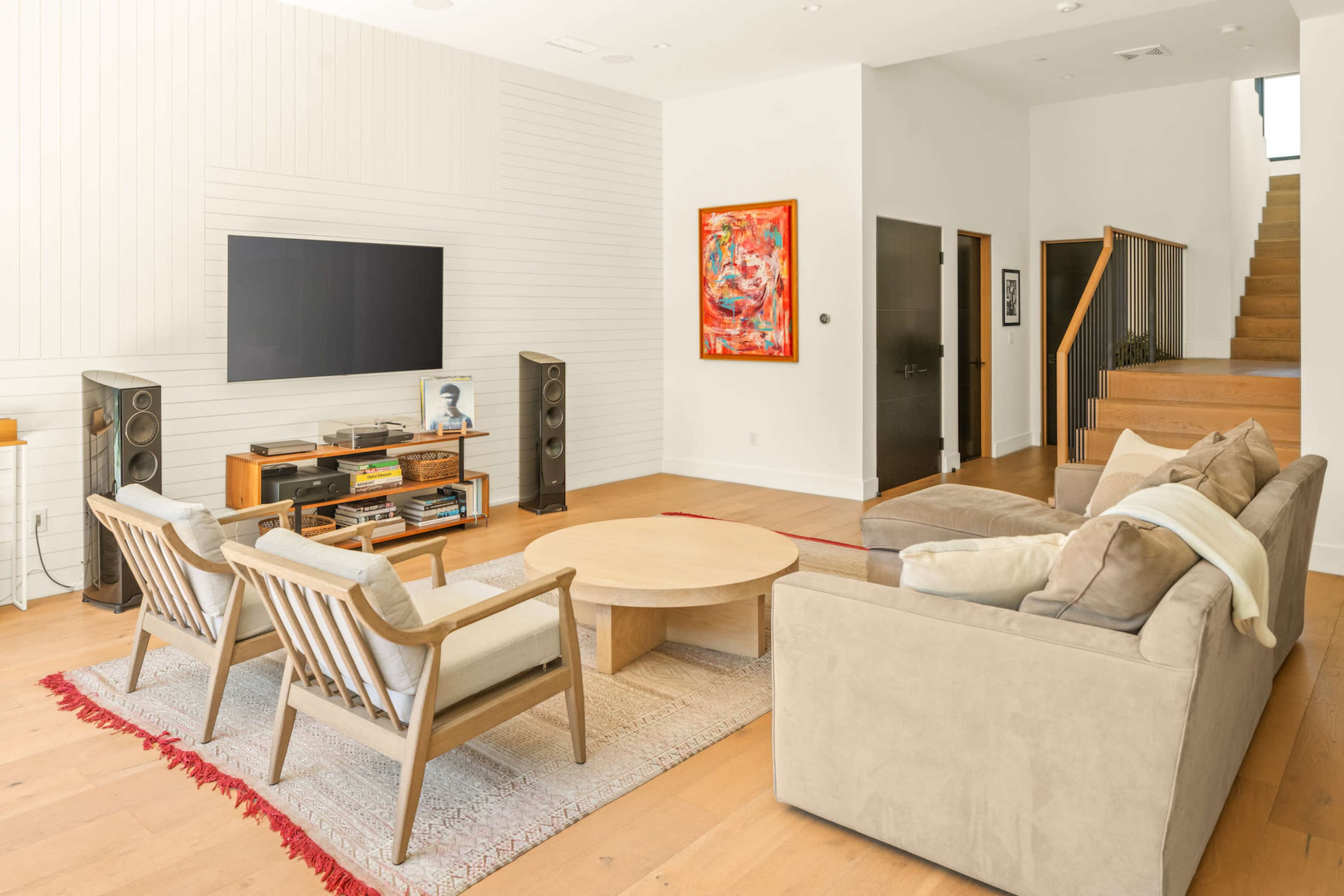 The living room features a large television mounted on the wall, a circular coffee table, two chairs, a sectional sofa, and a staircase leading to an upper level.