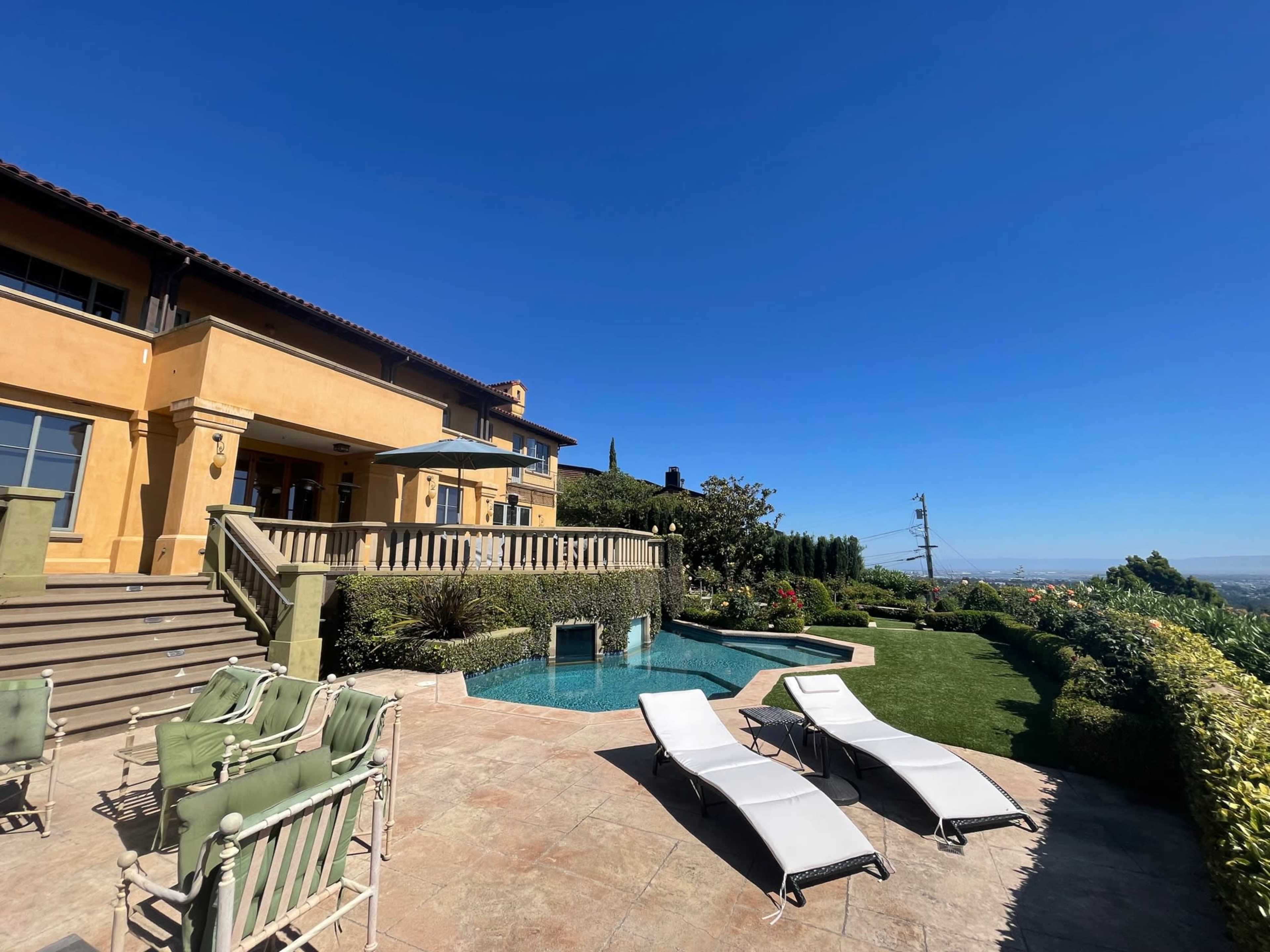 The image shows a spacious backyard of a house featuring a swimming pool, two lounge chairs, and well-maintained landscaping under a clear blue sky.