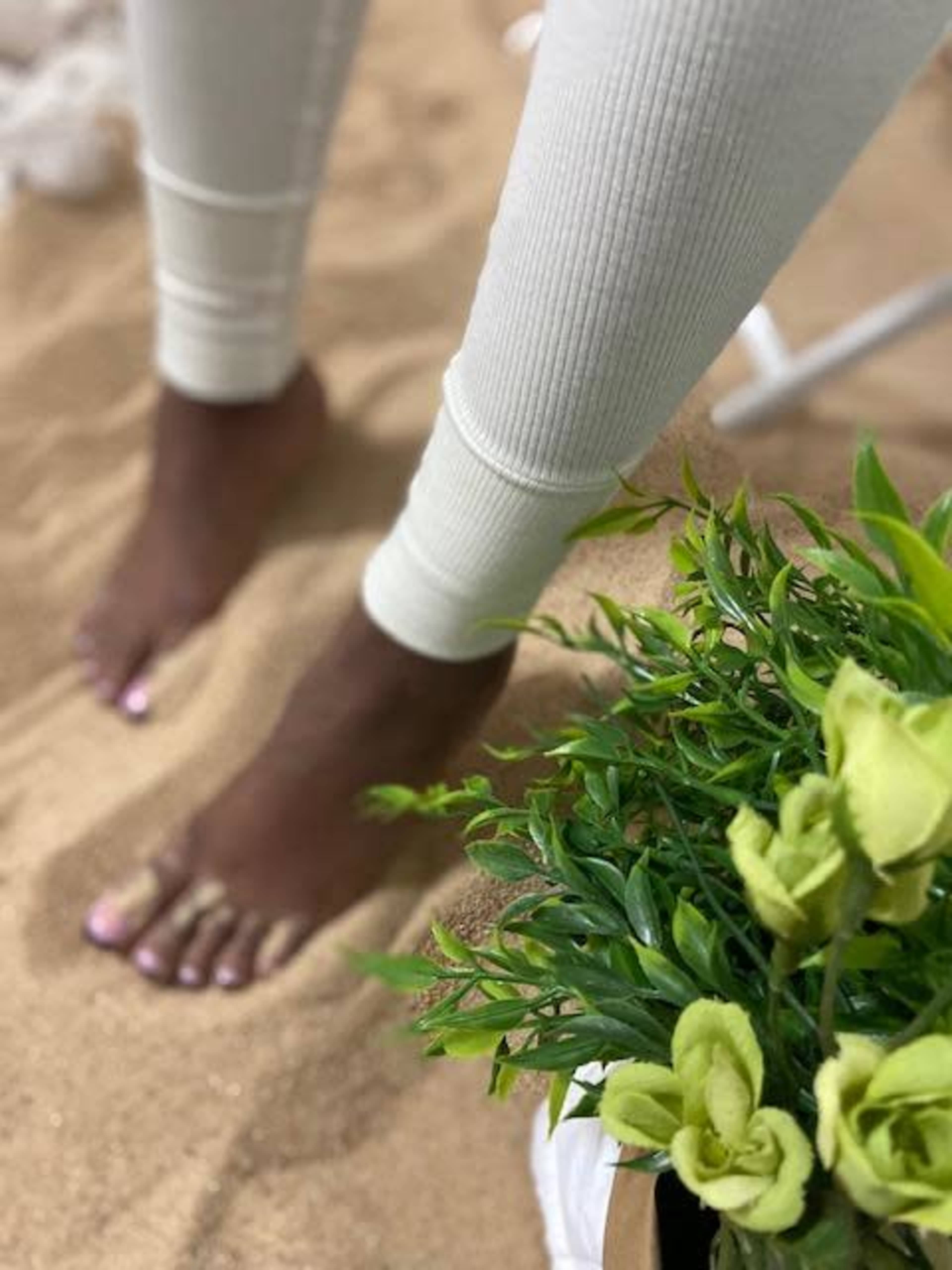 A person stands barefoot on sand next to a potted green plant.