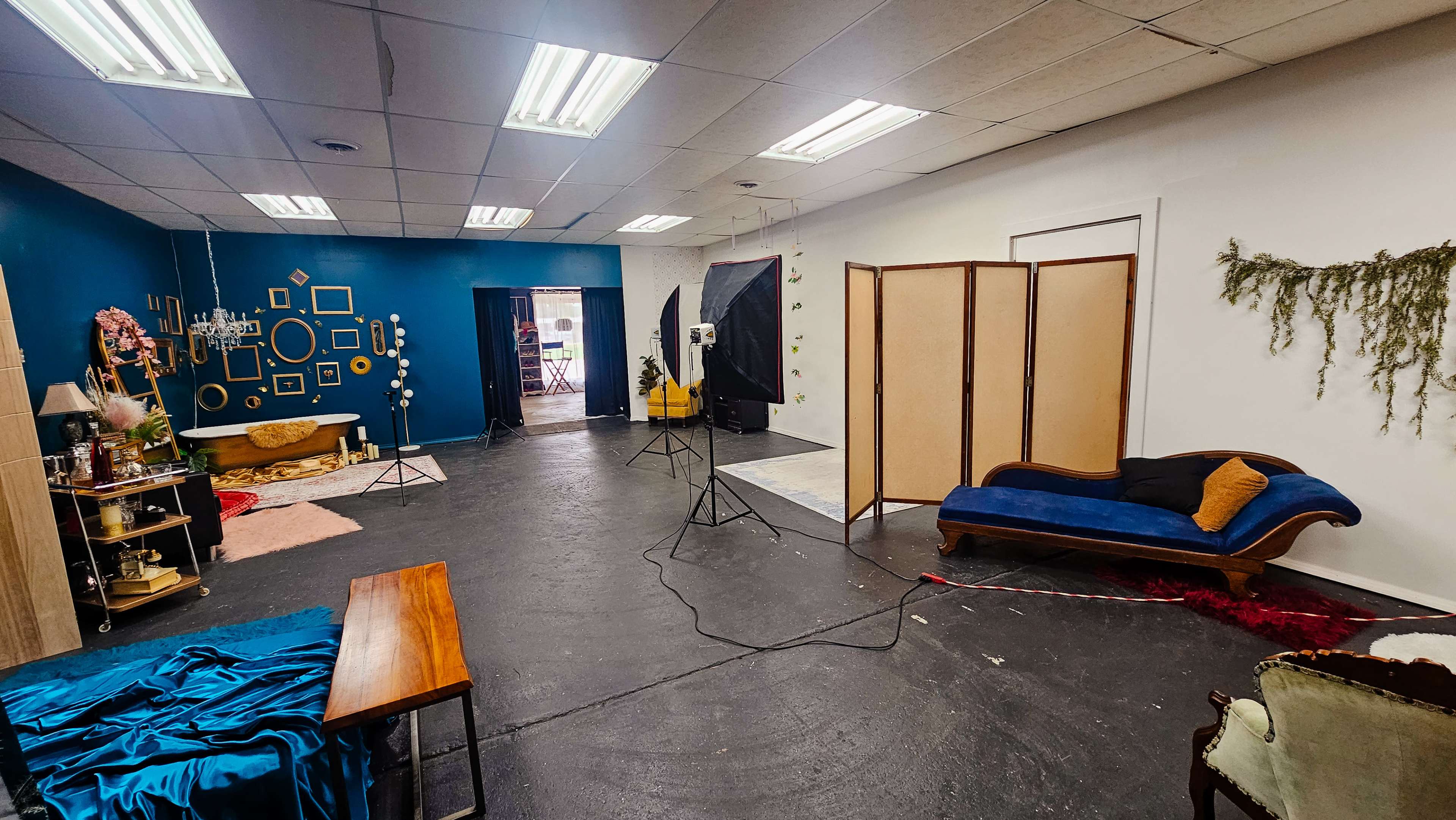 The image shows a spacious studio with various set components, including a blue accent wall, a vintage couch, photography equipment, and a folding screen.