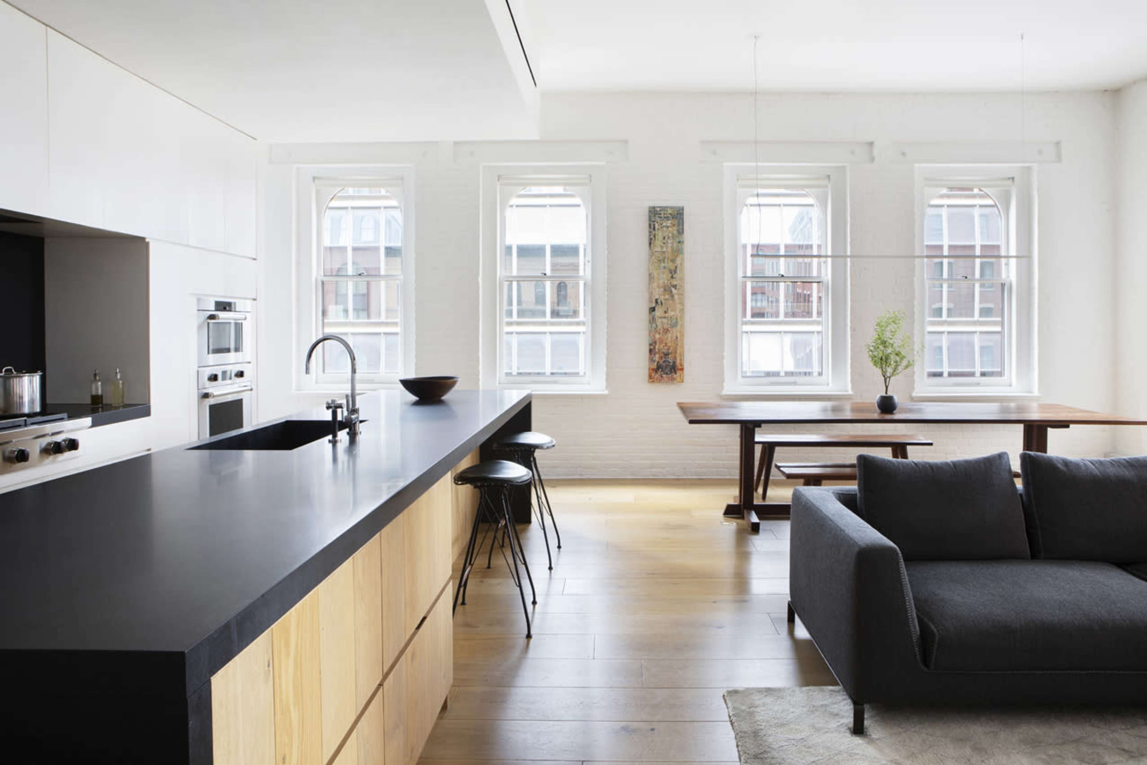 A modern kitchen and living area featuring a long black countertop, wooden cabinets, and large windows that illuminate the space.